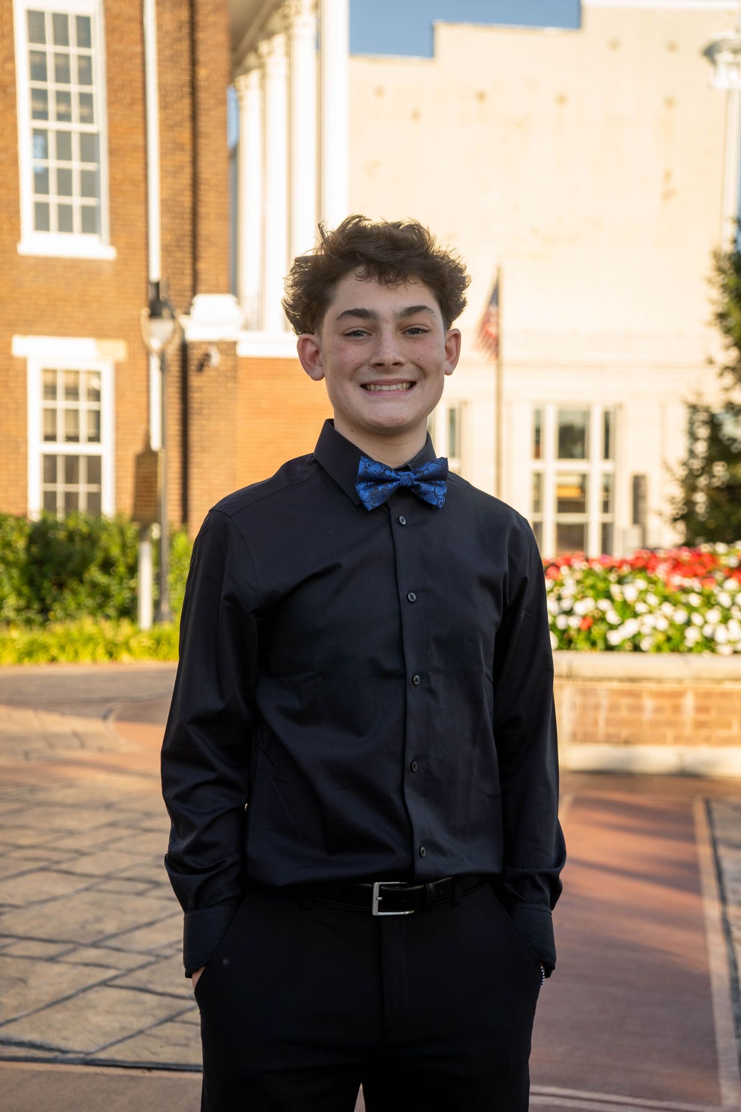 A smiling teenage boy in a dark shirt and blue bow tie poses outdoors before a formal dance.