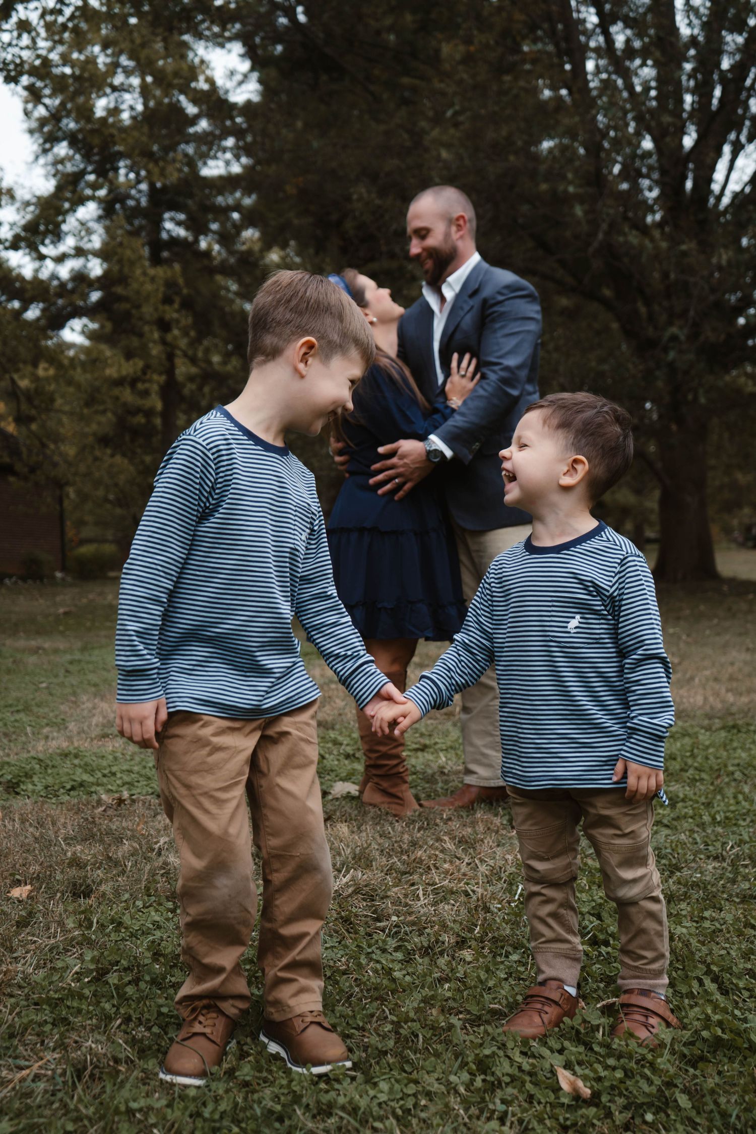 Two young brothers in matching striped shirts hold hands and smile while parents embrace romantically behind them outdoors.