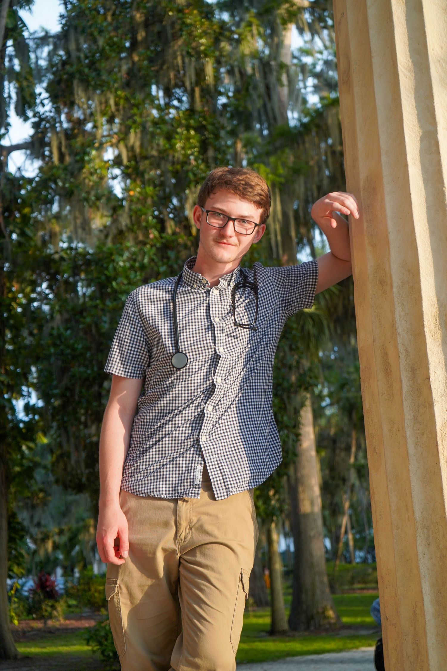 Young man in checkered shirt with stethoscope leaning against a column in a sunny outdoor park.