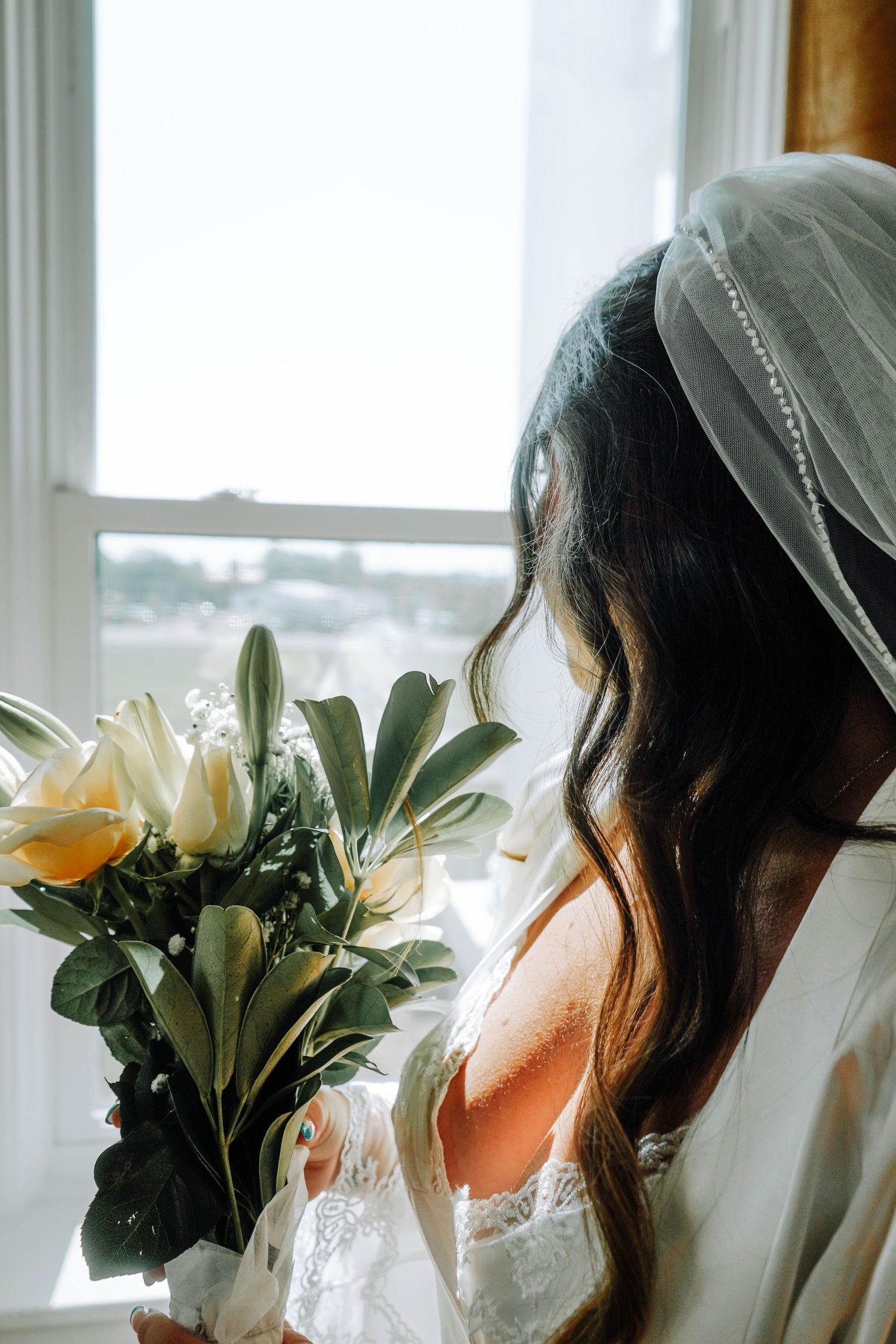 Bride wearing veil looks out sunlit window holding bouquet of white tulips, roses and greenery from behind.