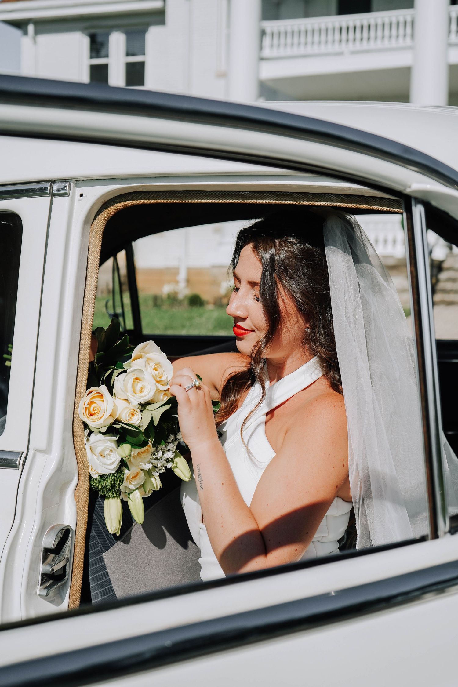 Bride in white halter wedding dress with red lips holds bouquet of white roses inside vintage classic car.