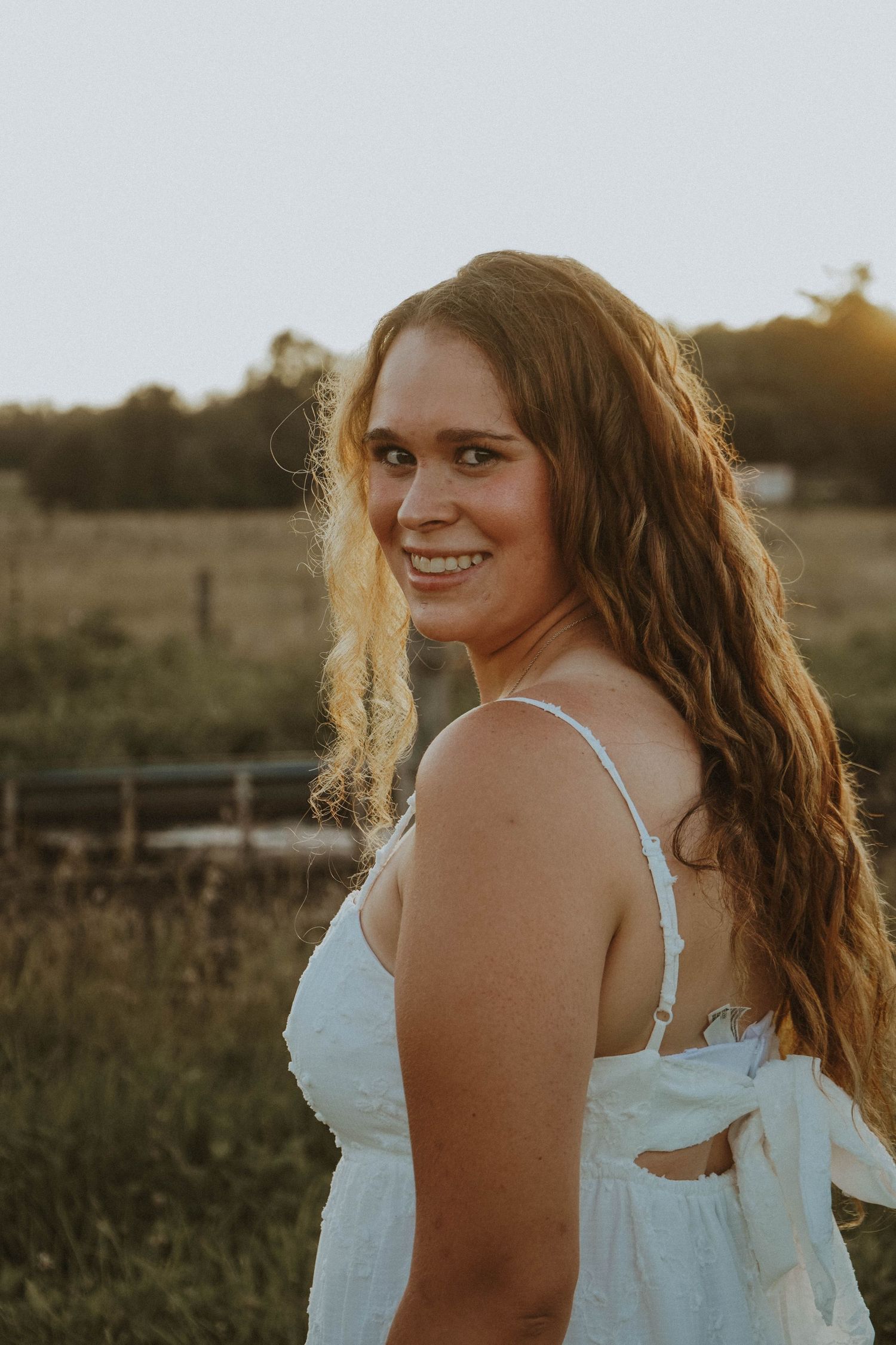 Smiling young woman with long wavy hair in white dress during golden hour outdoors.