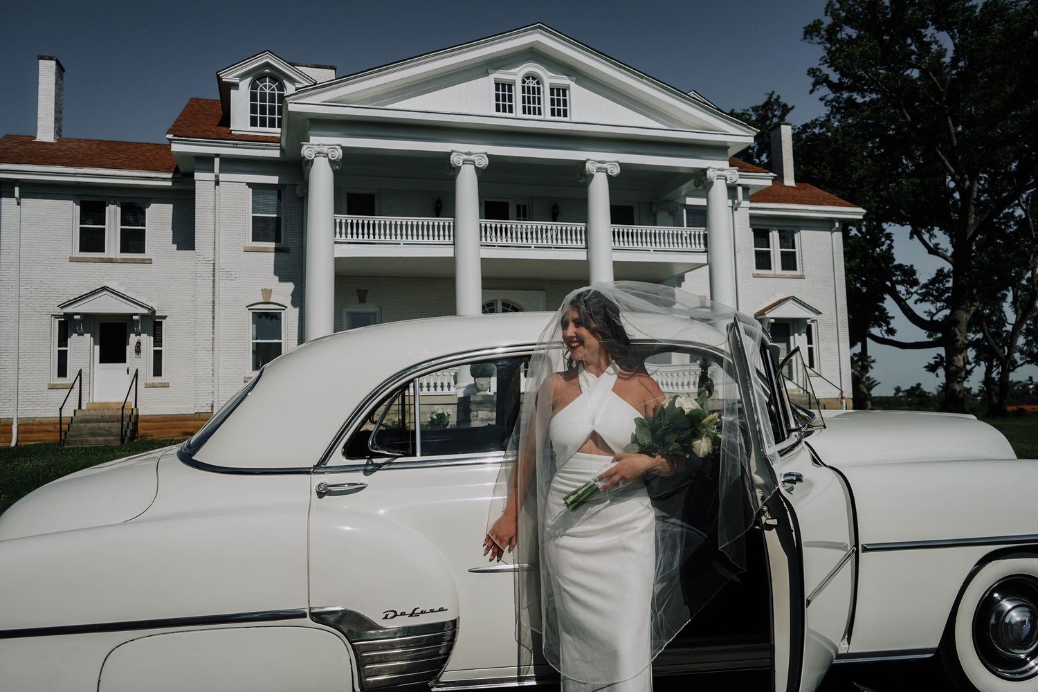 Bride in sleek white gown exiting a vintage Oldsmobile classic car before a grand white colonial estate.