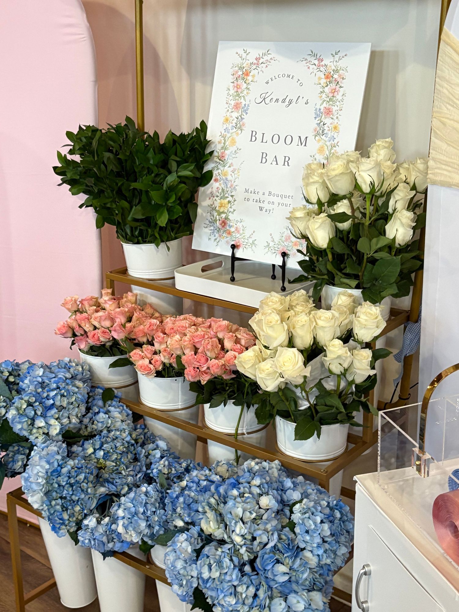 A bloom bar display with white roses, pink flowers, and blue hydrangeas arranged in white pots on a gold shelf.