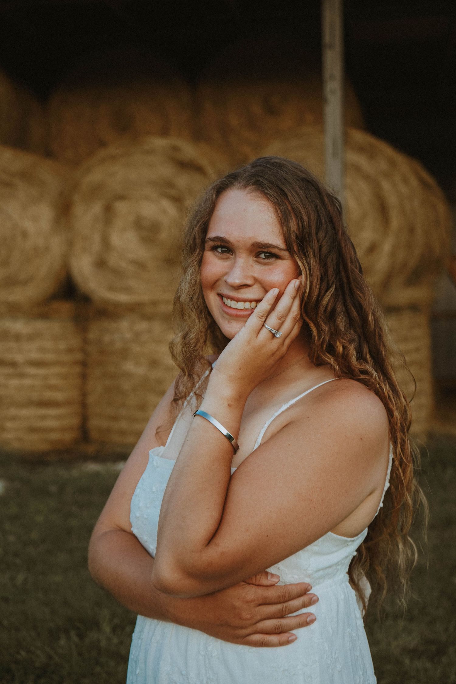 Smiling young woman in white sundress posing in front of hay bales on a farm.