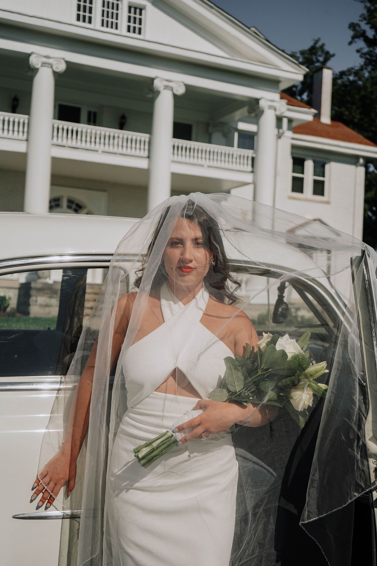 Bride in modern white halter gown with flowing veil holds bouquet while exiting vintage car at estate venue.