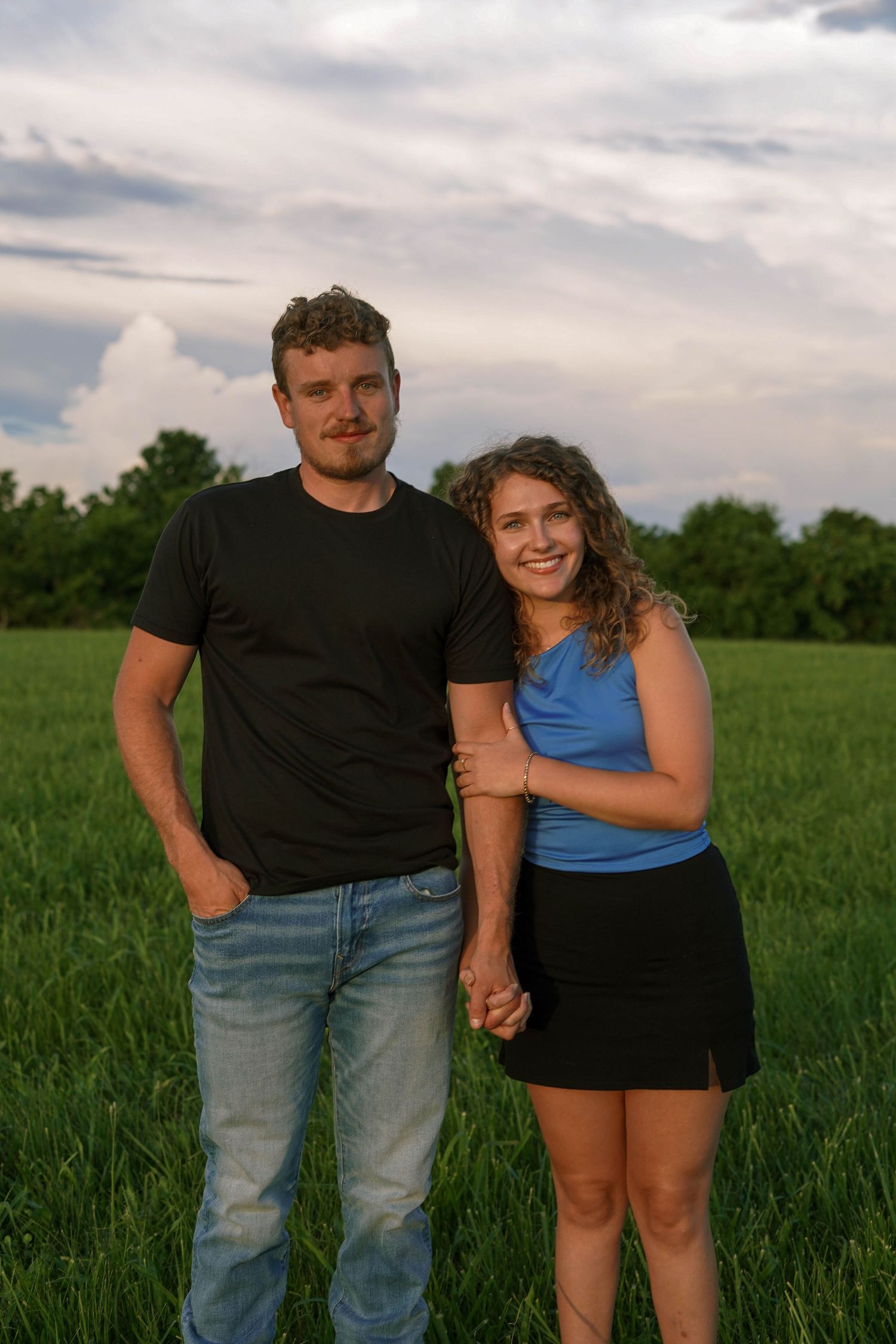 Happy couple holding hands in a lush green field under dramatic cloudy sky at dusk.