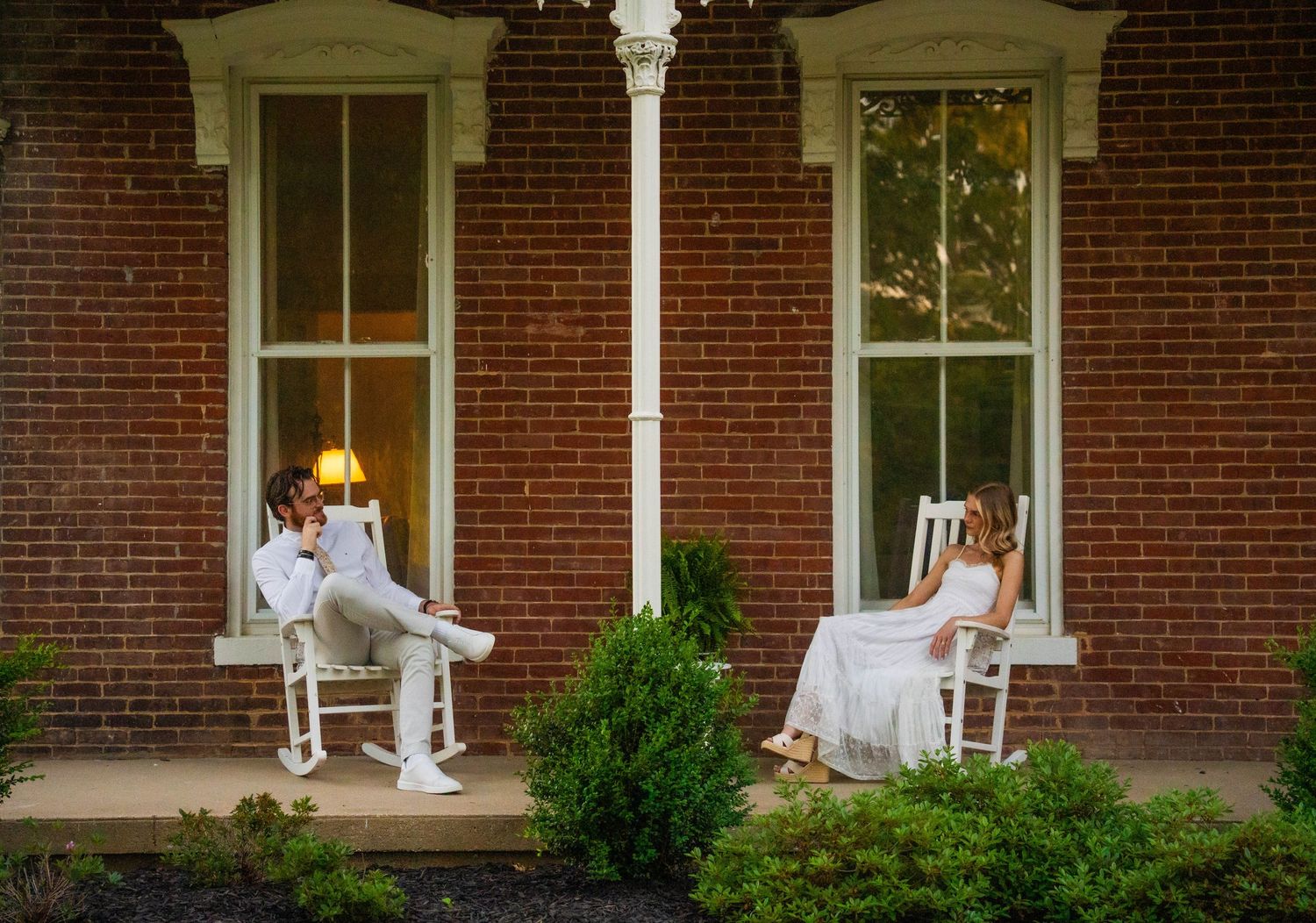 Couple in wedding attire sitting in white rocking chairs on a brick house porch, sharing a romantic moment.
