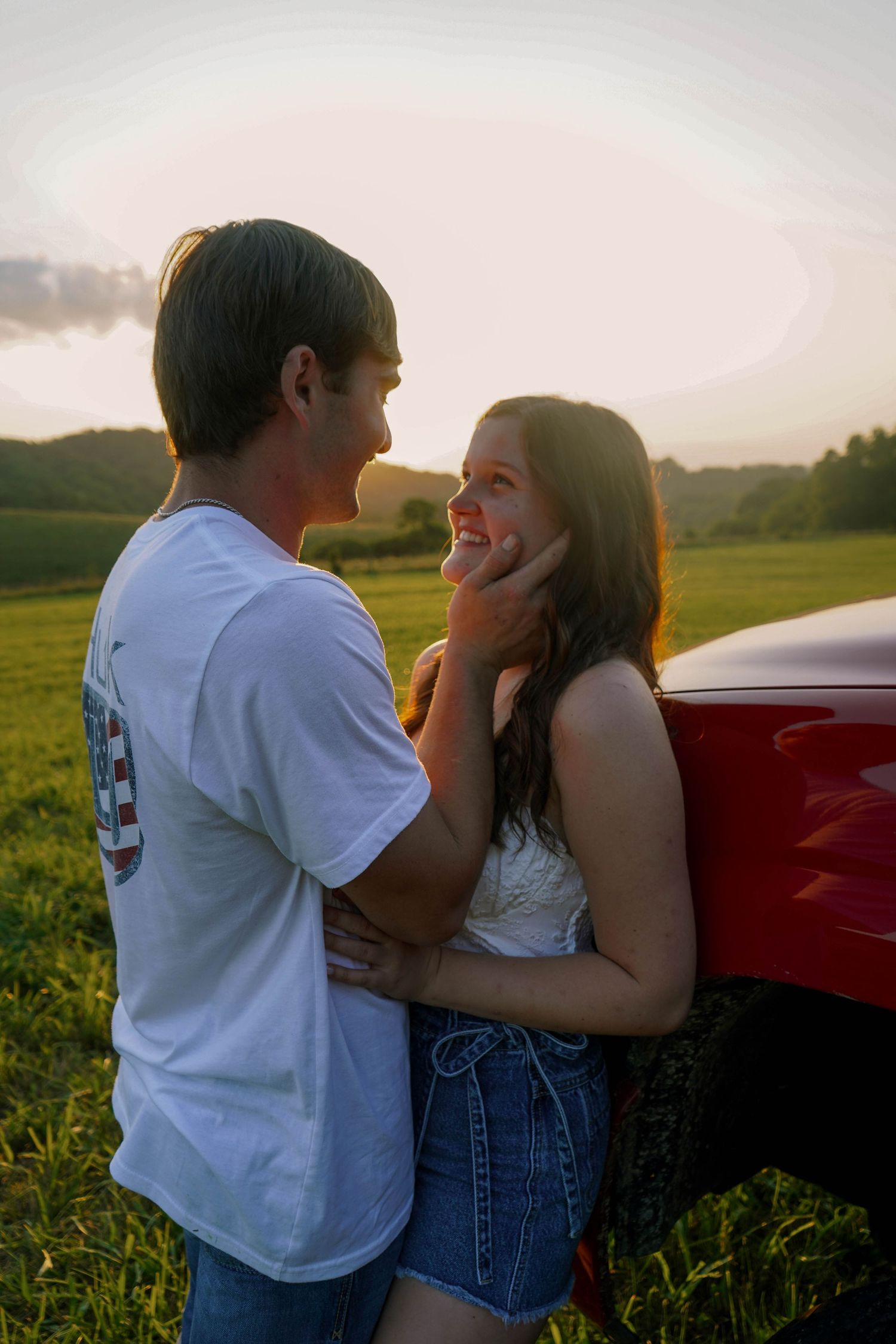 Young couple gazing at each other tenderly at sunset, man gently holding womans face near a red pickup truck in a field.