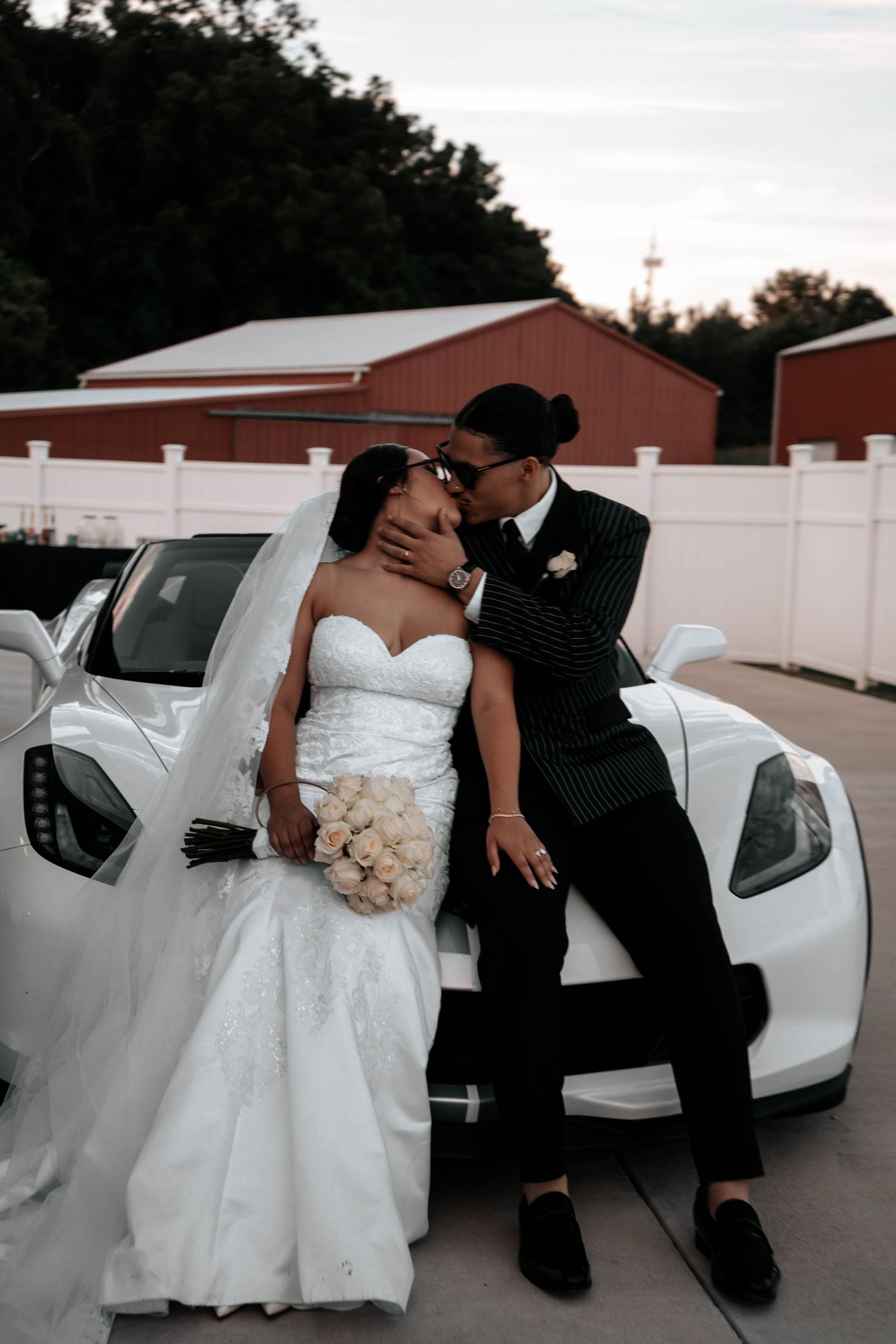 Bride in strapless lace gown and groom in pinstripe suit share a tender kiss leaning on white Corvette.