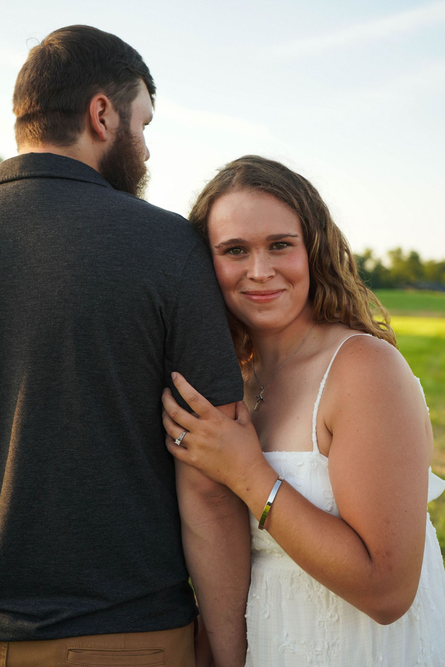Smiling woman in white dress leaning on bearded mans shoulder during outdoor engagement session.
