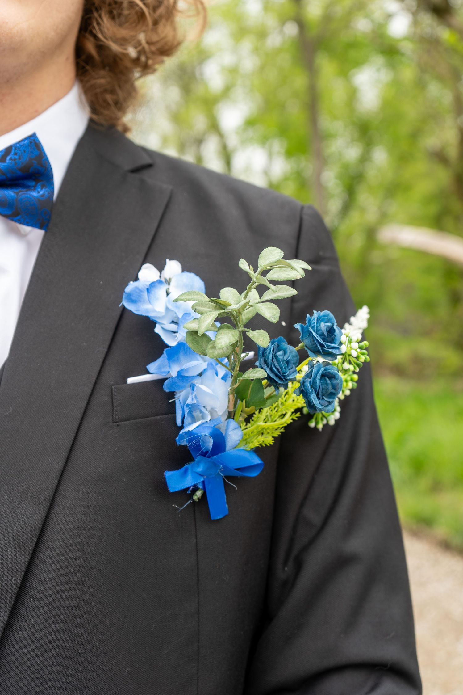Close-up of a blue floral boutonniere with roses and greenery on a black tuxedo lapel with blue polka dot bow tie.