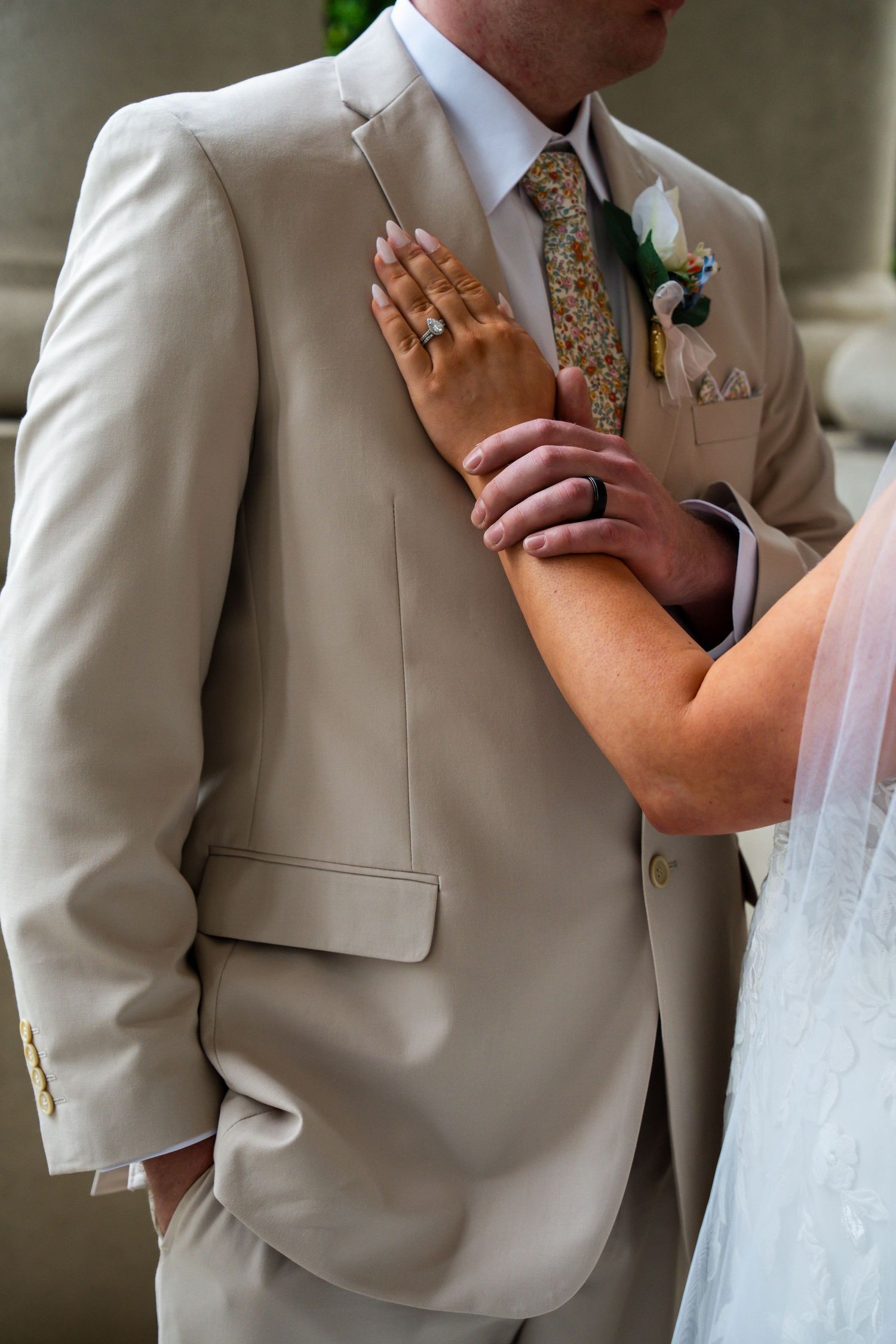 Bride places her hand on groom's tan suit lapel, showing off wedding ring, with floral boutonniere visible.