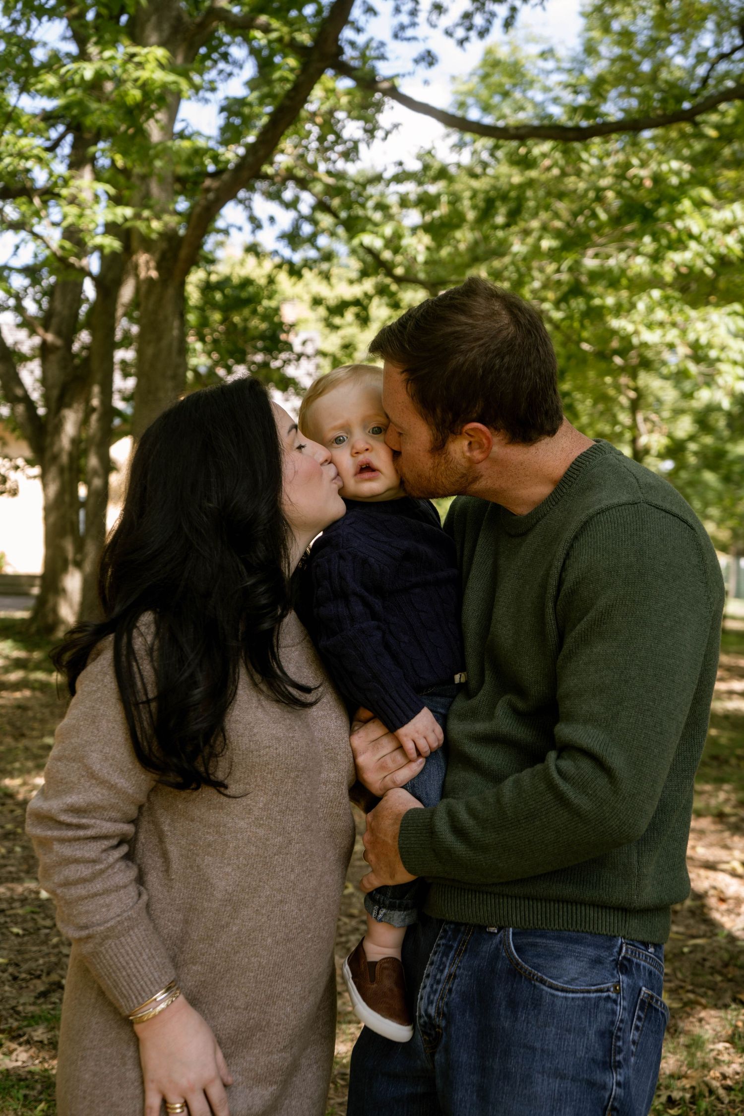Loving parents kissing their toddler on both cheeks simultaneously in a lush outdoor park setting.
