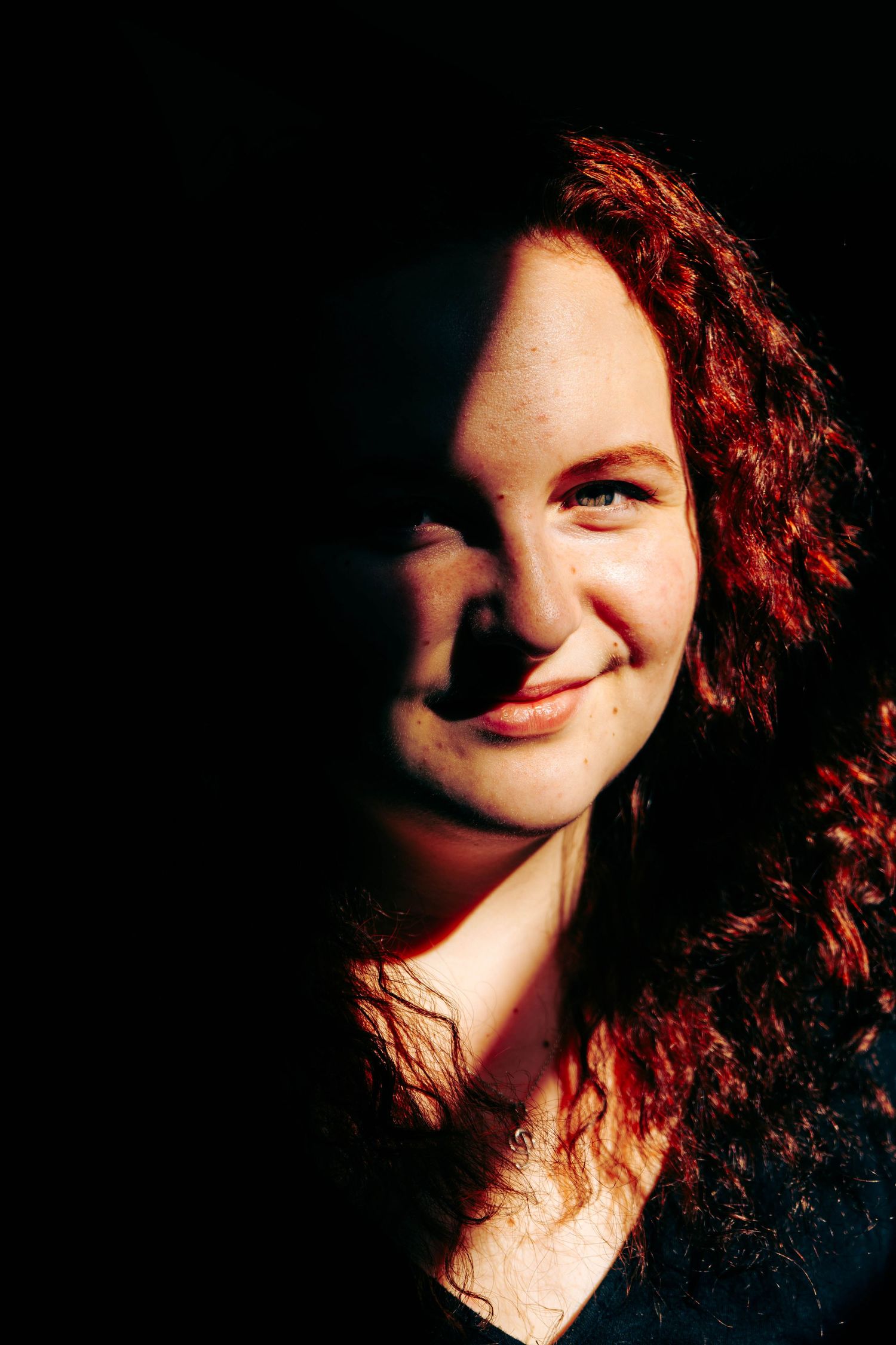 Smiling young woman with curly red hair illuminated by dramatic side lighting against dark background.