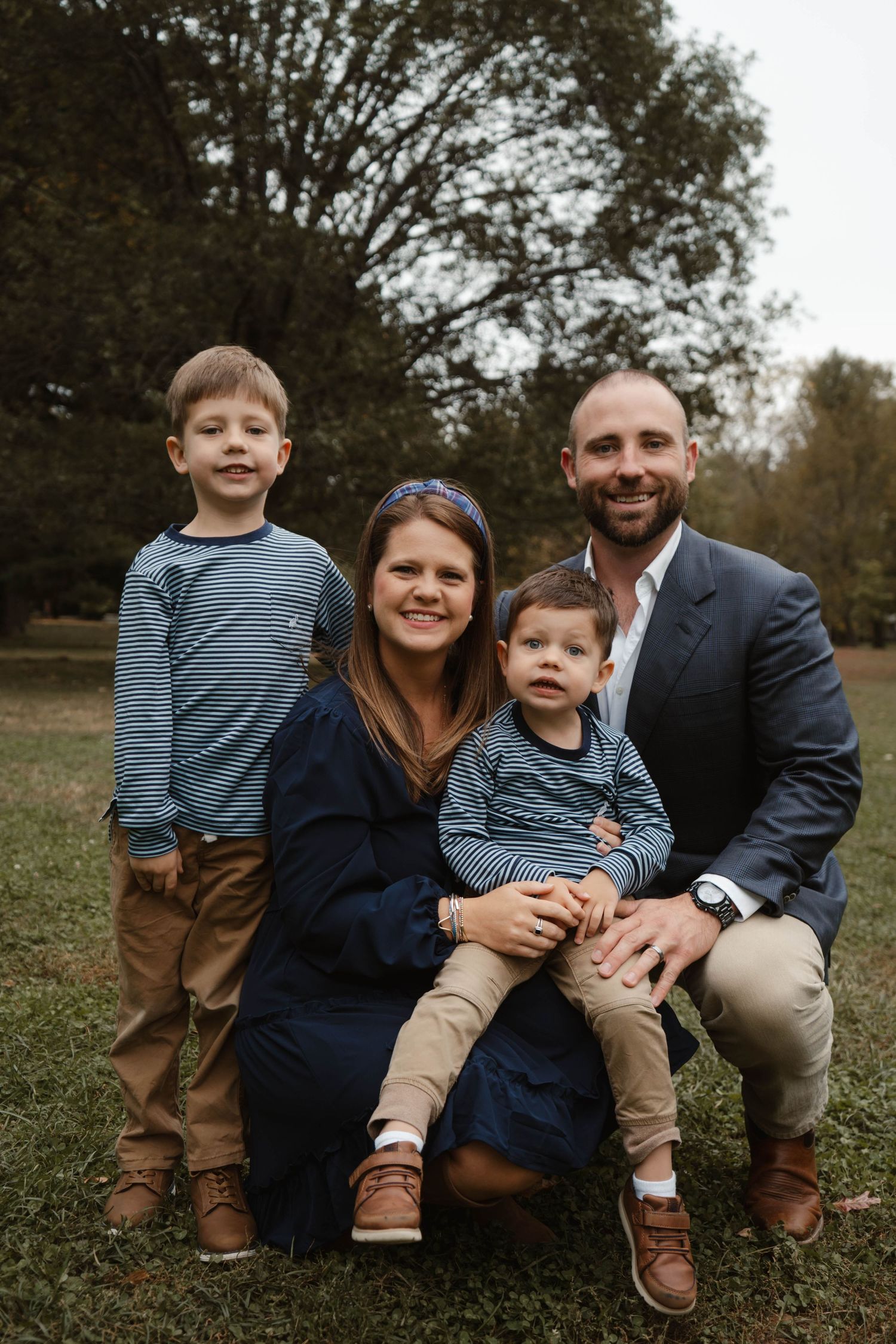 Family of four poses in park, mother kneeling with toddler on lap while father and older son stand beside them.