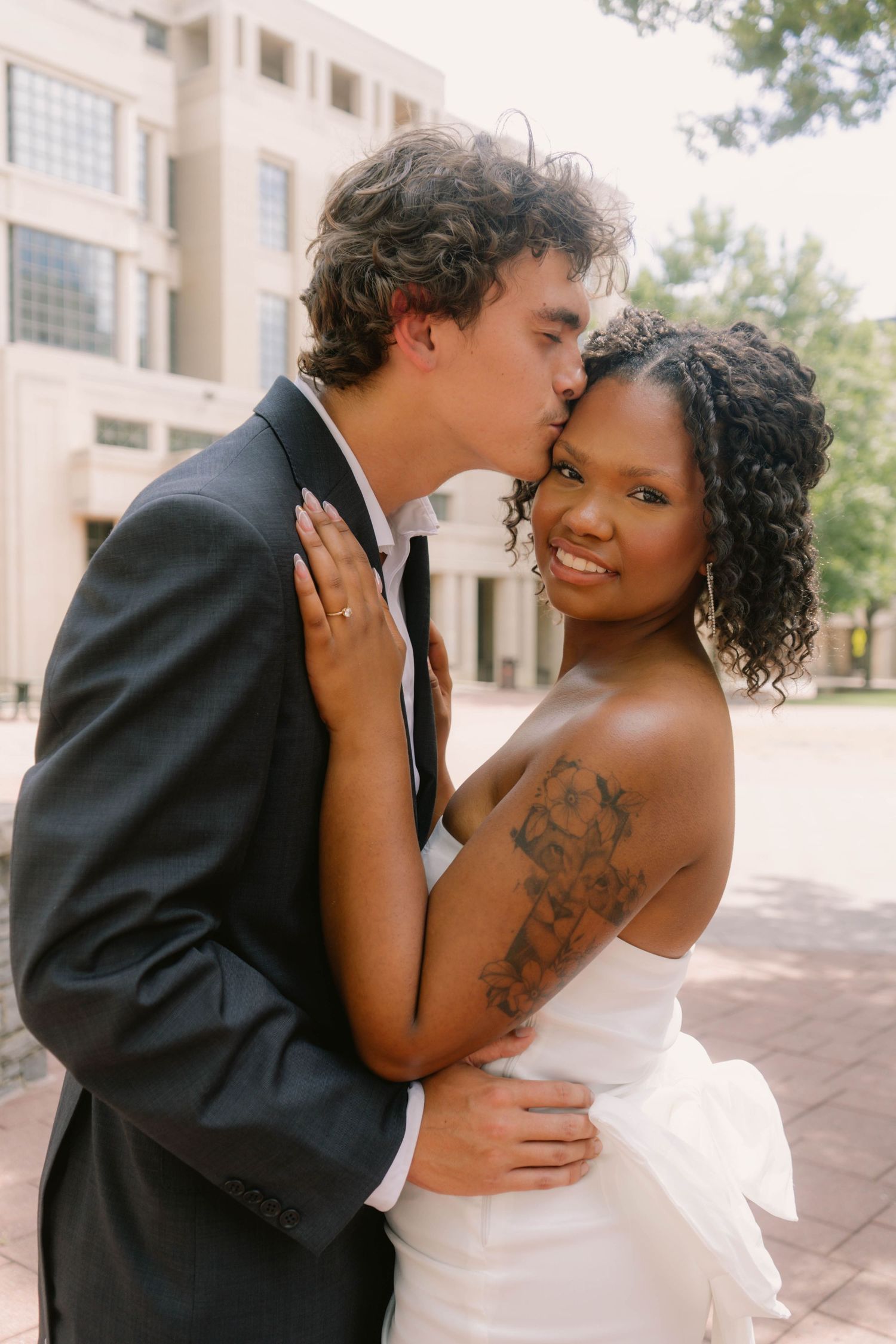 Groom kissing bride on cheek outdoors as tattooed bride in strapless white dress smiles at camera.