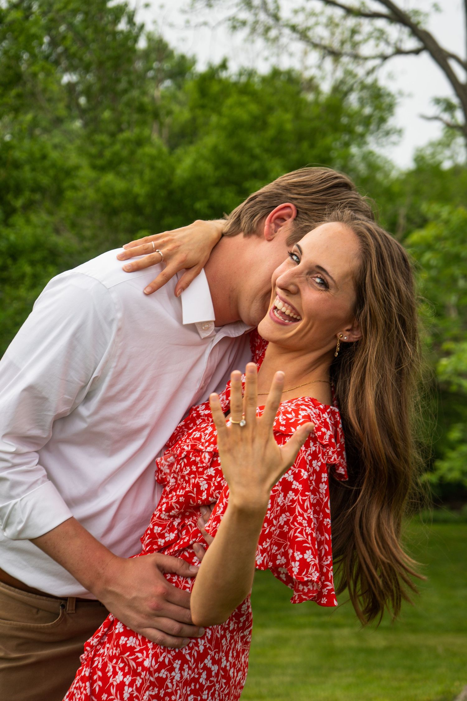 Joyful woman in red floral dress shows off engagement ring while man embraces her from behind outdoors.