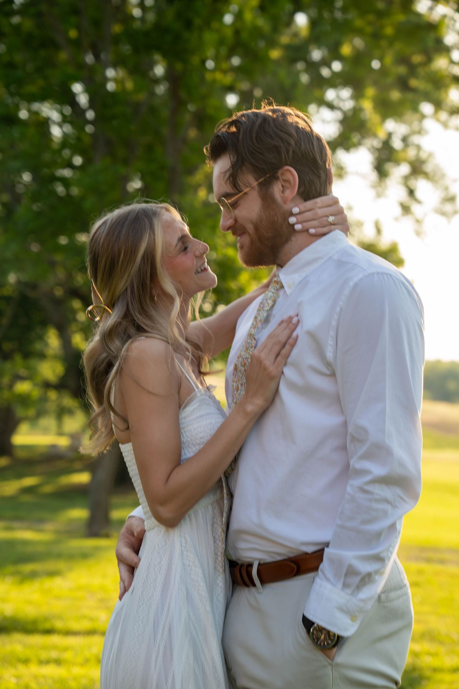 Couple in white wedding attire embrace and smile at each other in a sunlit outdoor garden setting.