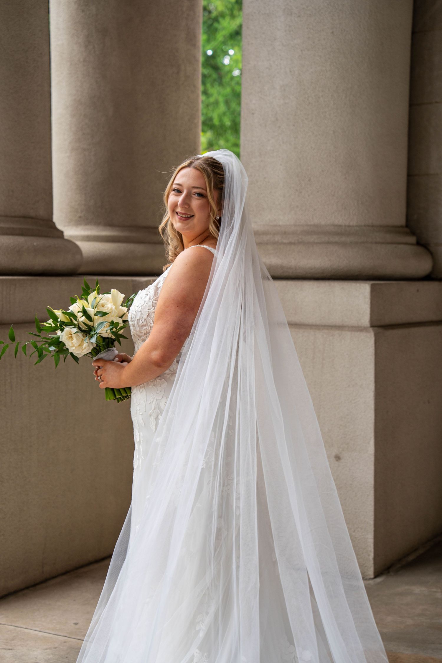 Smiling bride in lace gown and long veil holds white rose bouquet between stone columns.