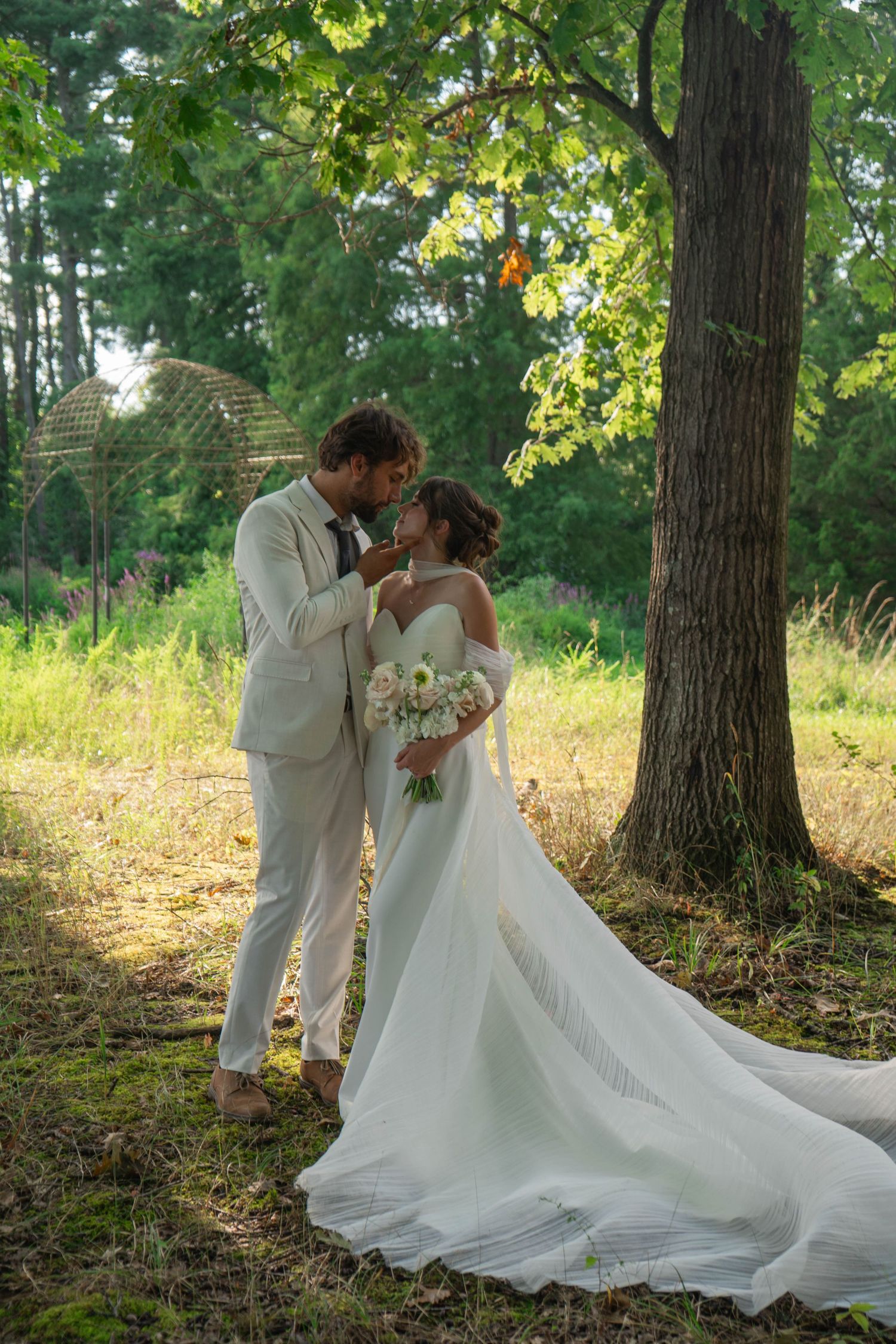 Bride and groom standing intimately under a tree in golden sunlit garden setting.