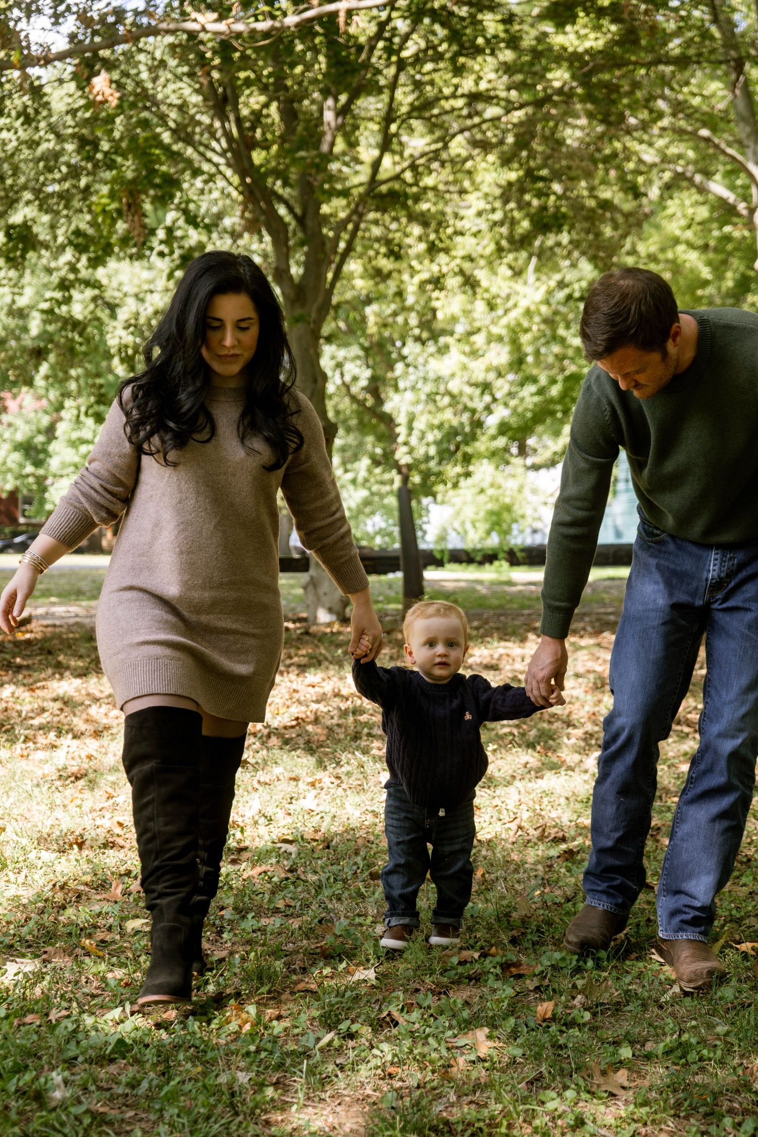 Young family helping their toddler walk through a green park, parents holding his hands on each side.
