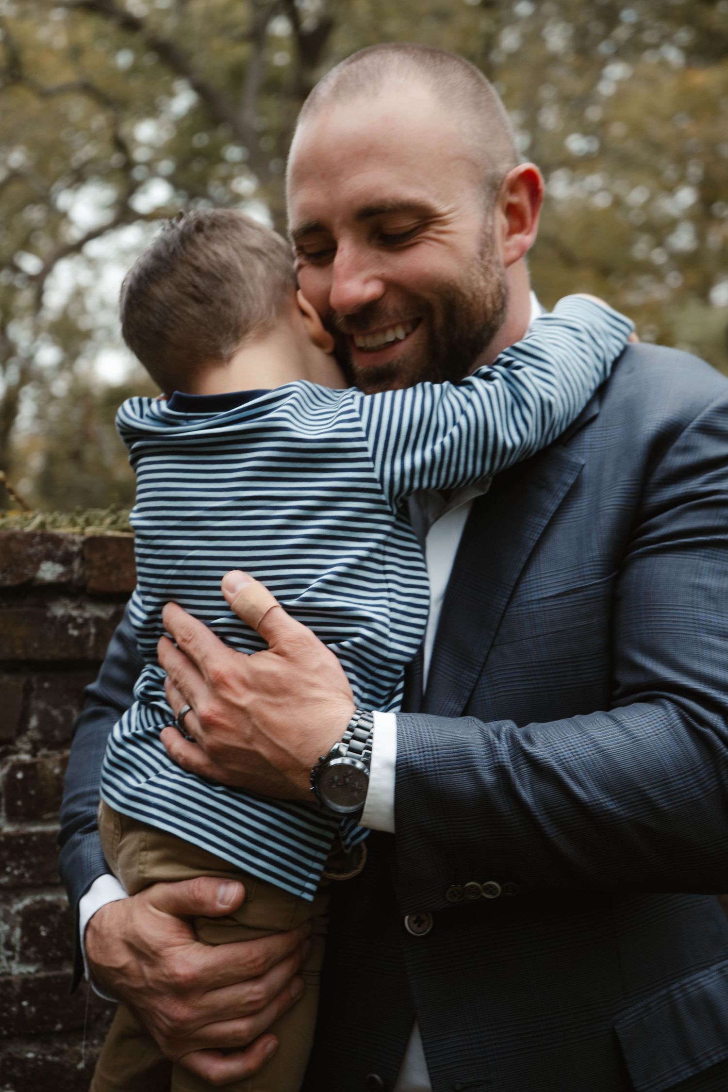 Smiling bald father in navy blazer tenderly hugs his young toddler son outdoors near a brick wall.