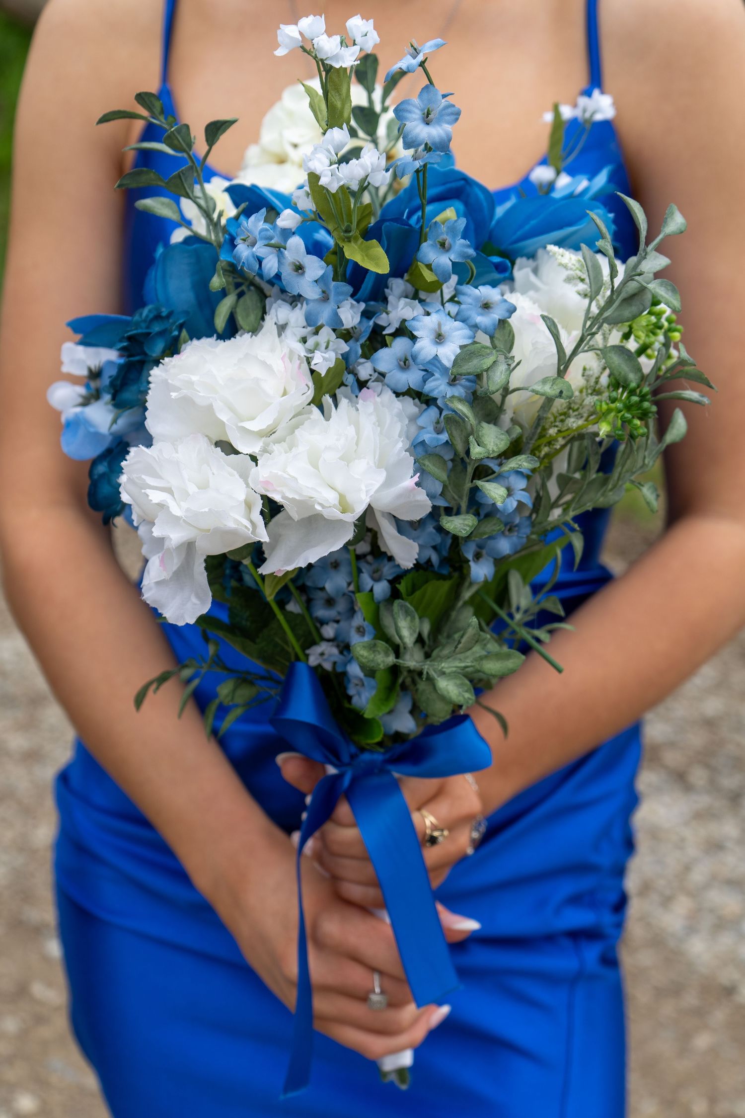 Close-up of a blue and white prom bouquet with roses and wildflowers tied with royal blue ribbon, held by woman in blue dress.
