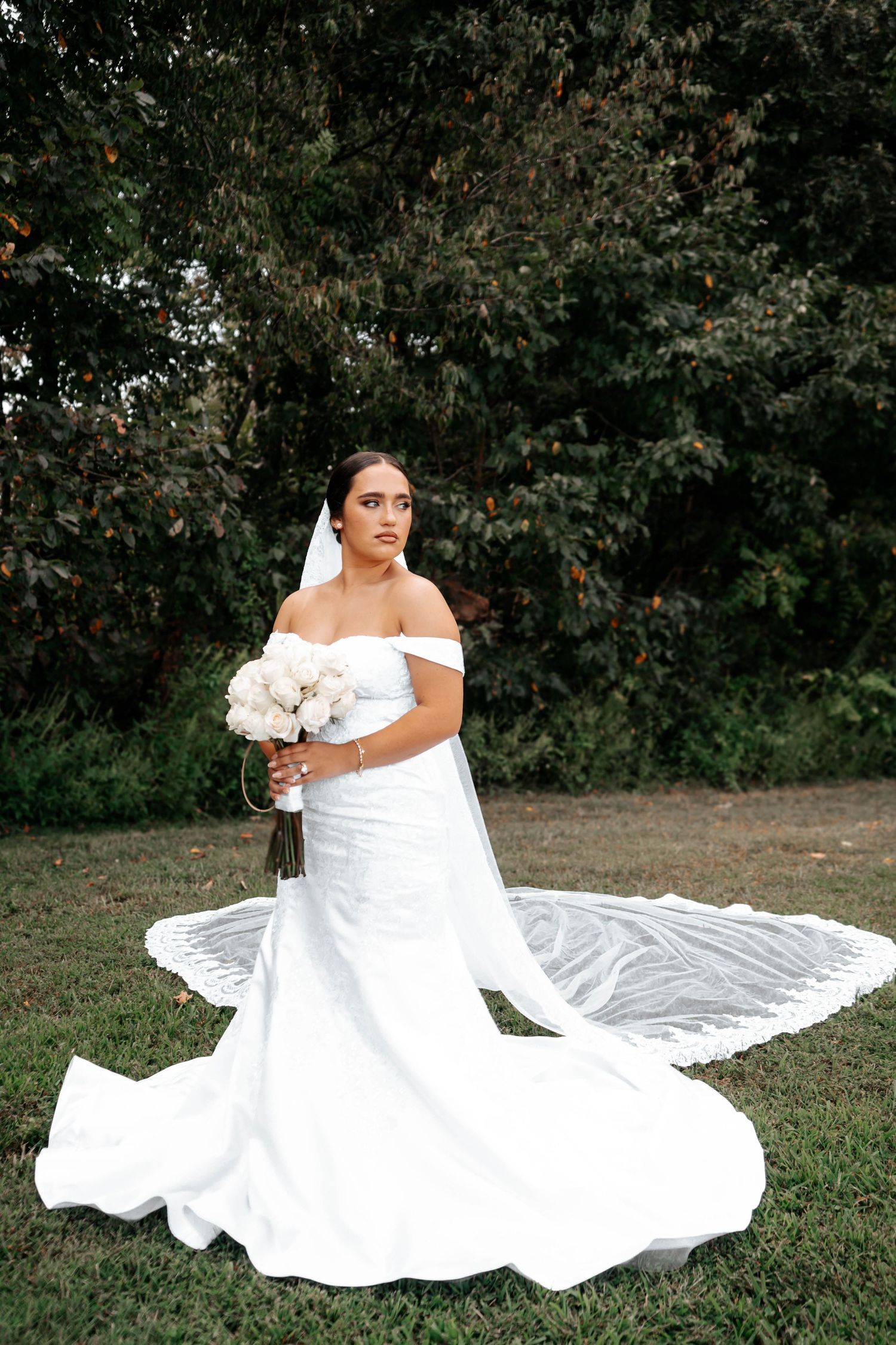 Bride in off-shoulder mermaid gown poses with white bouquet on lush green lawn.