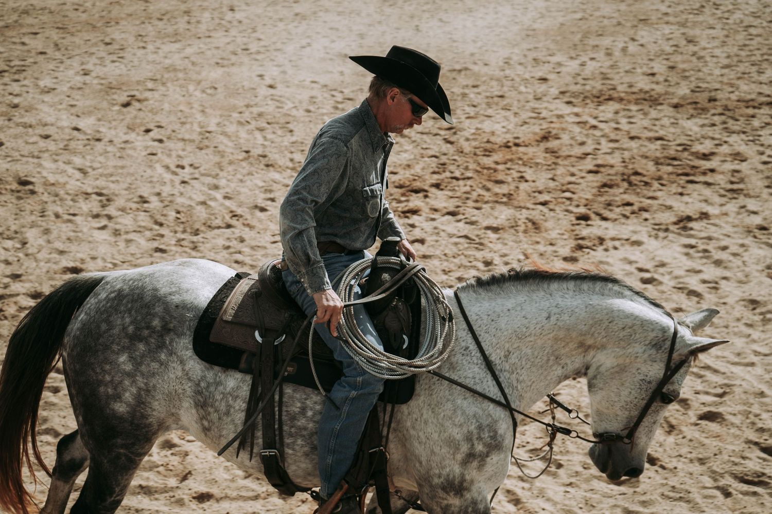 Cowboy riding a dapple gray horse while holding a lasso in a sandy rodeo arena.
