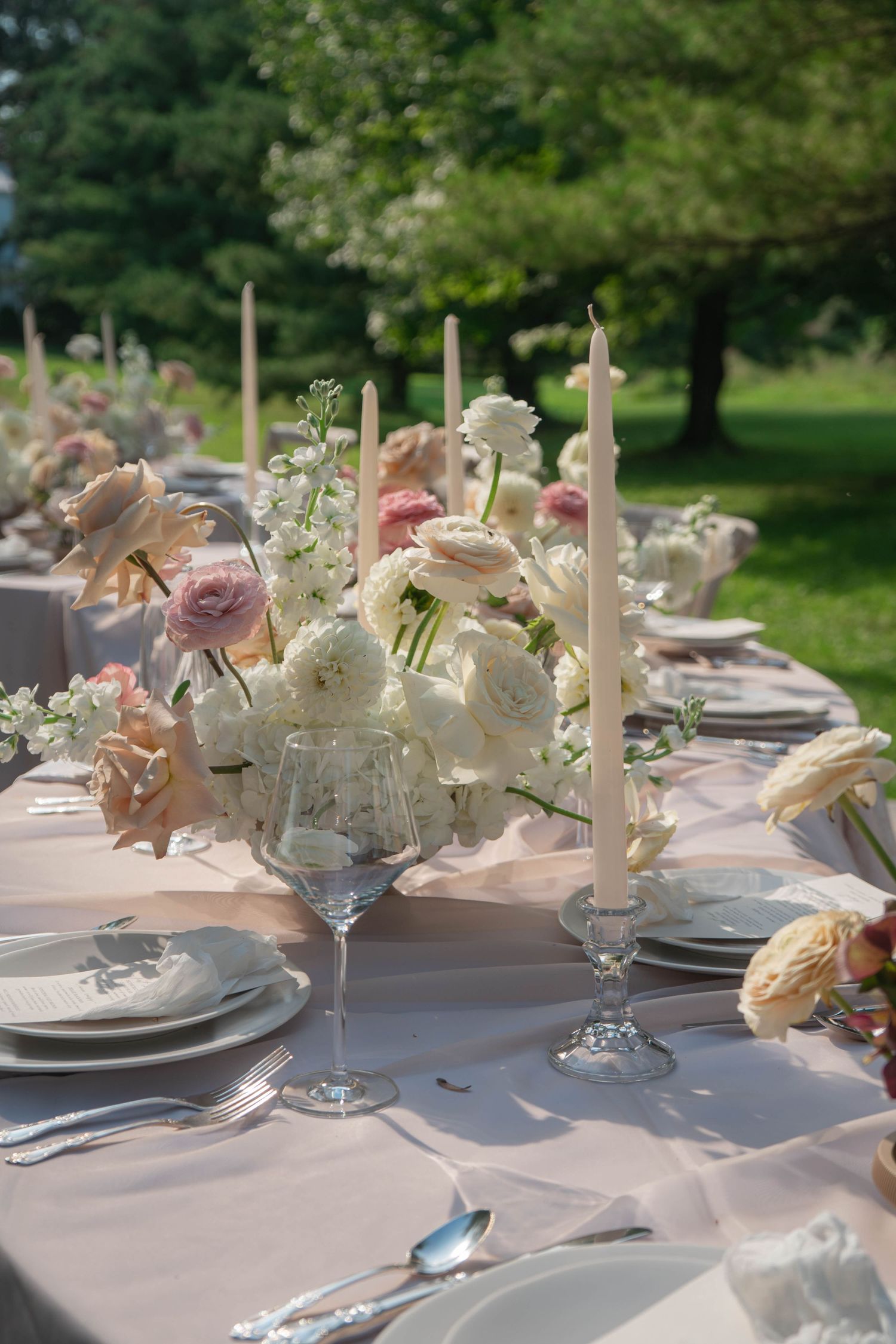 Outdoor wedding reception table with blush linens, white floral centerpieces, and tall taper candles.