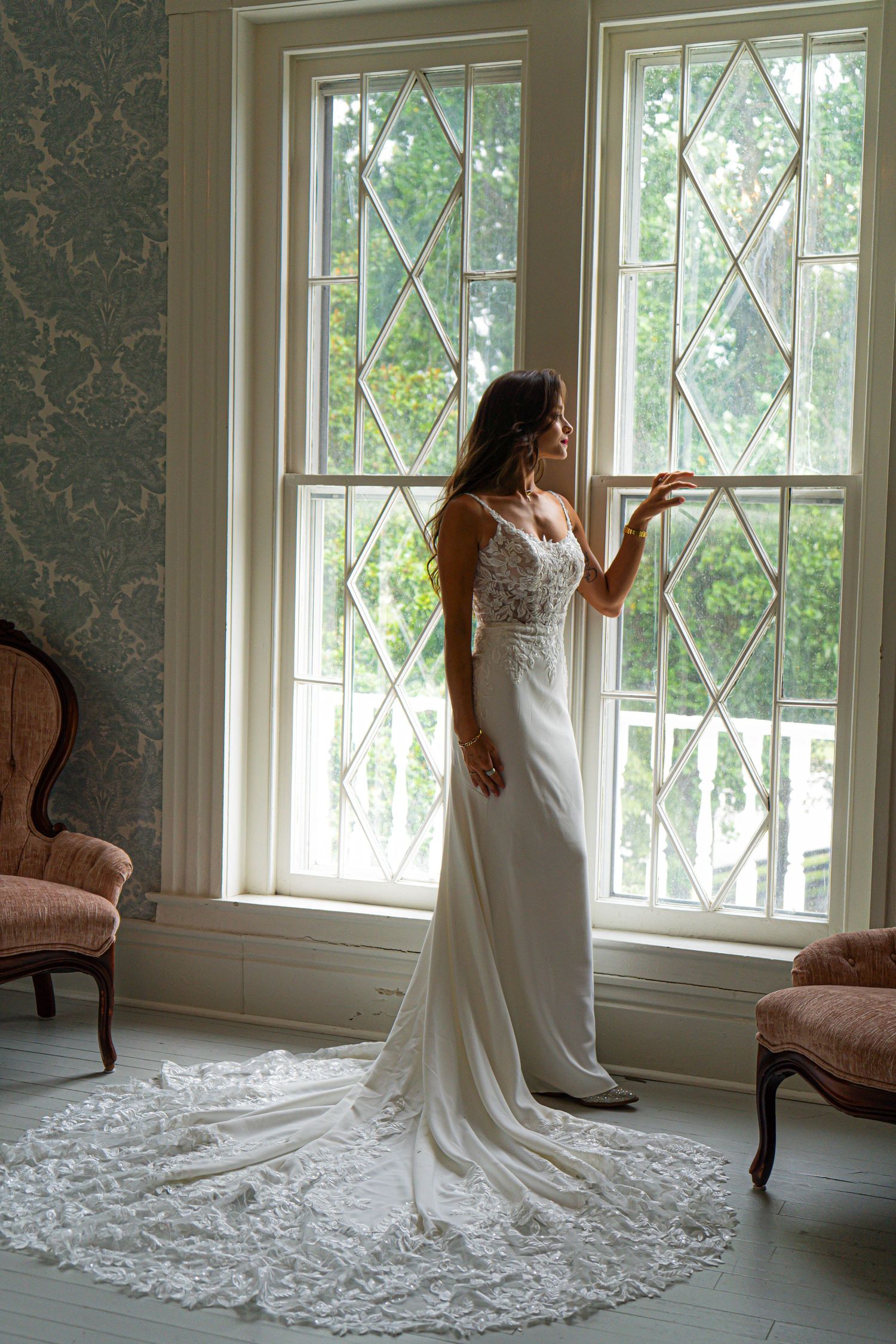 Bride in elegant lace wedding gown with train stands by diamond-paned windows in a vintage room with pink chairs.