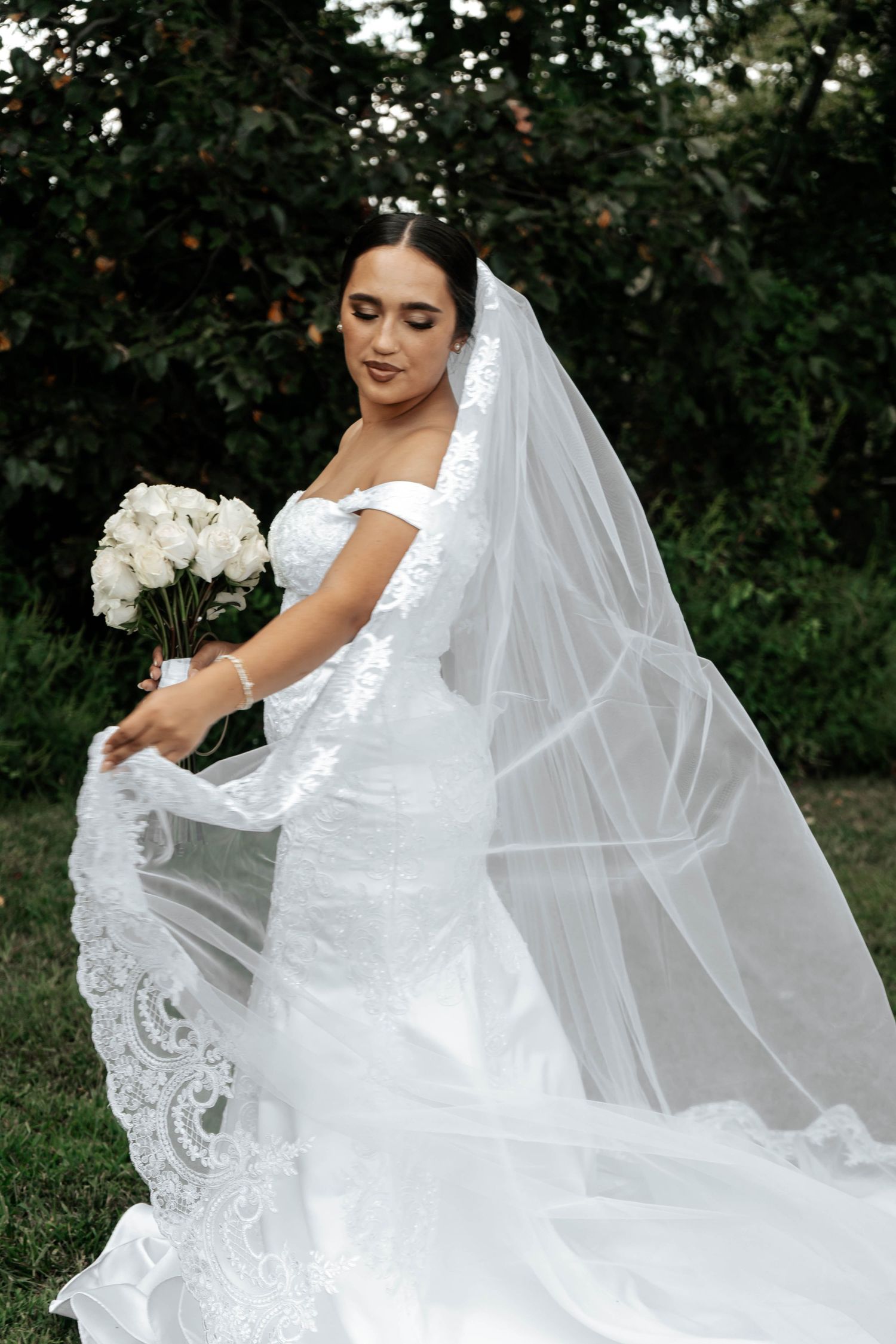 Beautiful bride in lace mermaid gown displays her long veil while holding white rose bouquet outdoors.