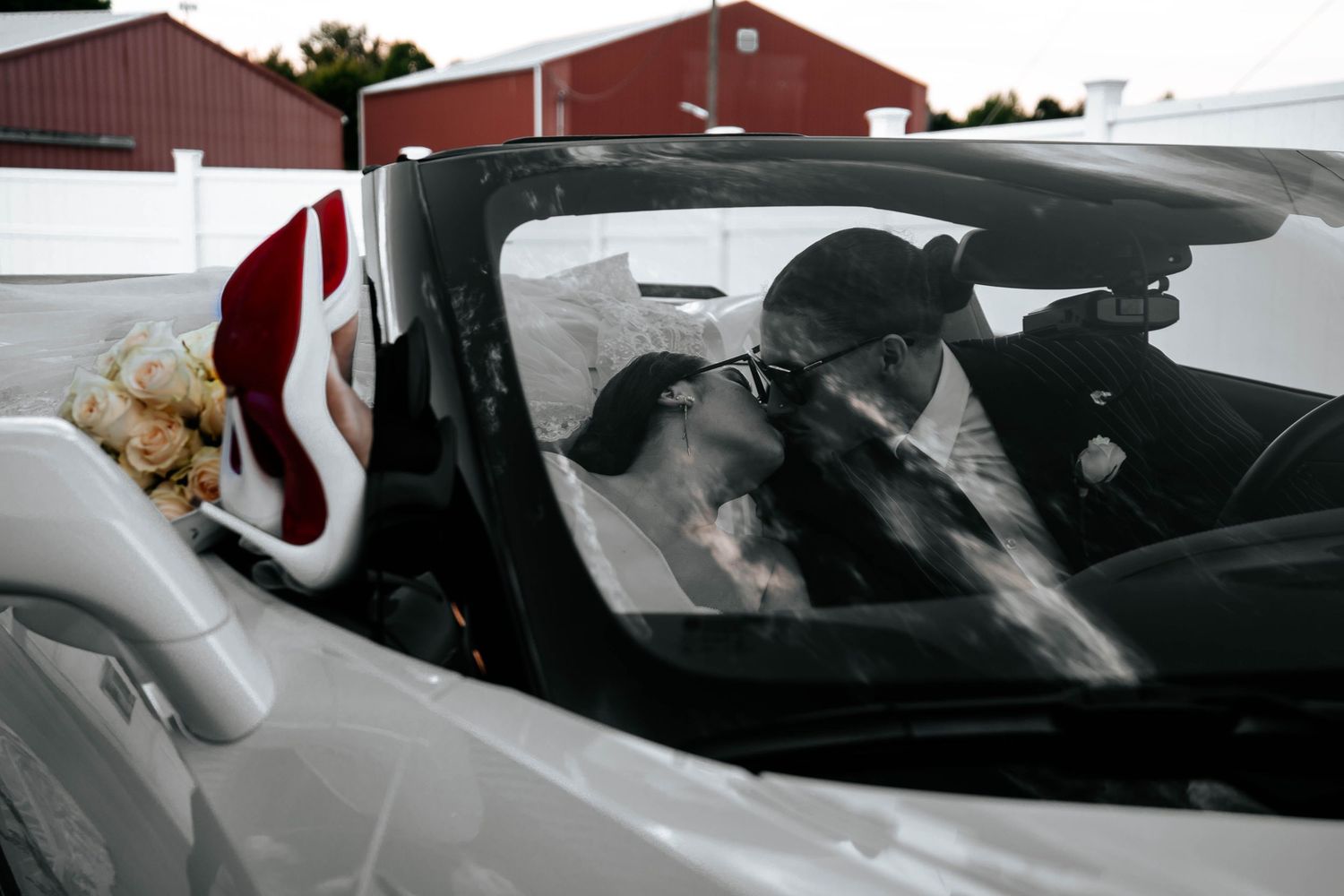 Newlyweds share a kiss inside a white convertible car with bridal bouquet visible through the windshield.