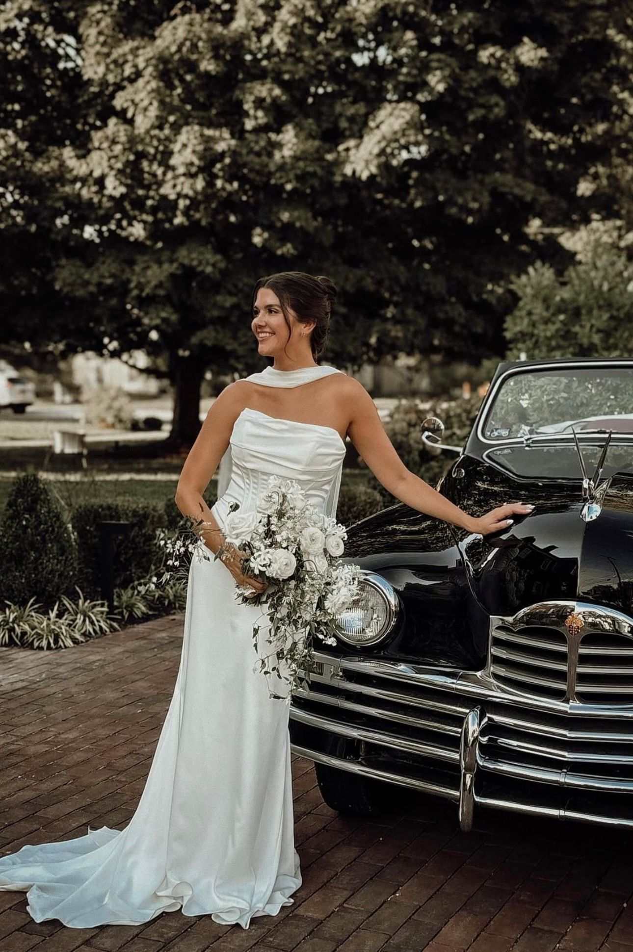 Elegant bride in sleek white gown poses with vintage black car holding white floral bouquet.