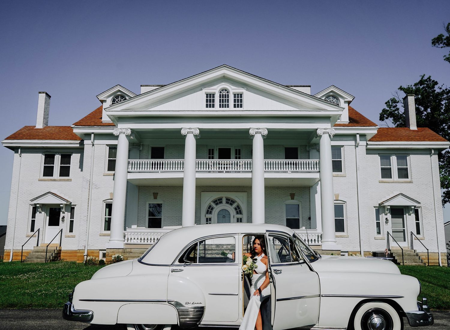 Bride and groom posing with vintage white car in front of elegant white colonial mansion.
