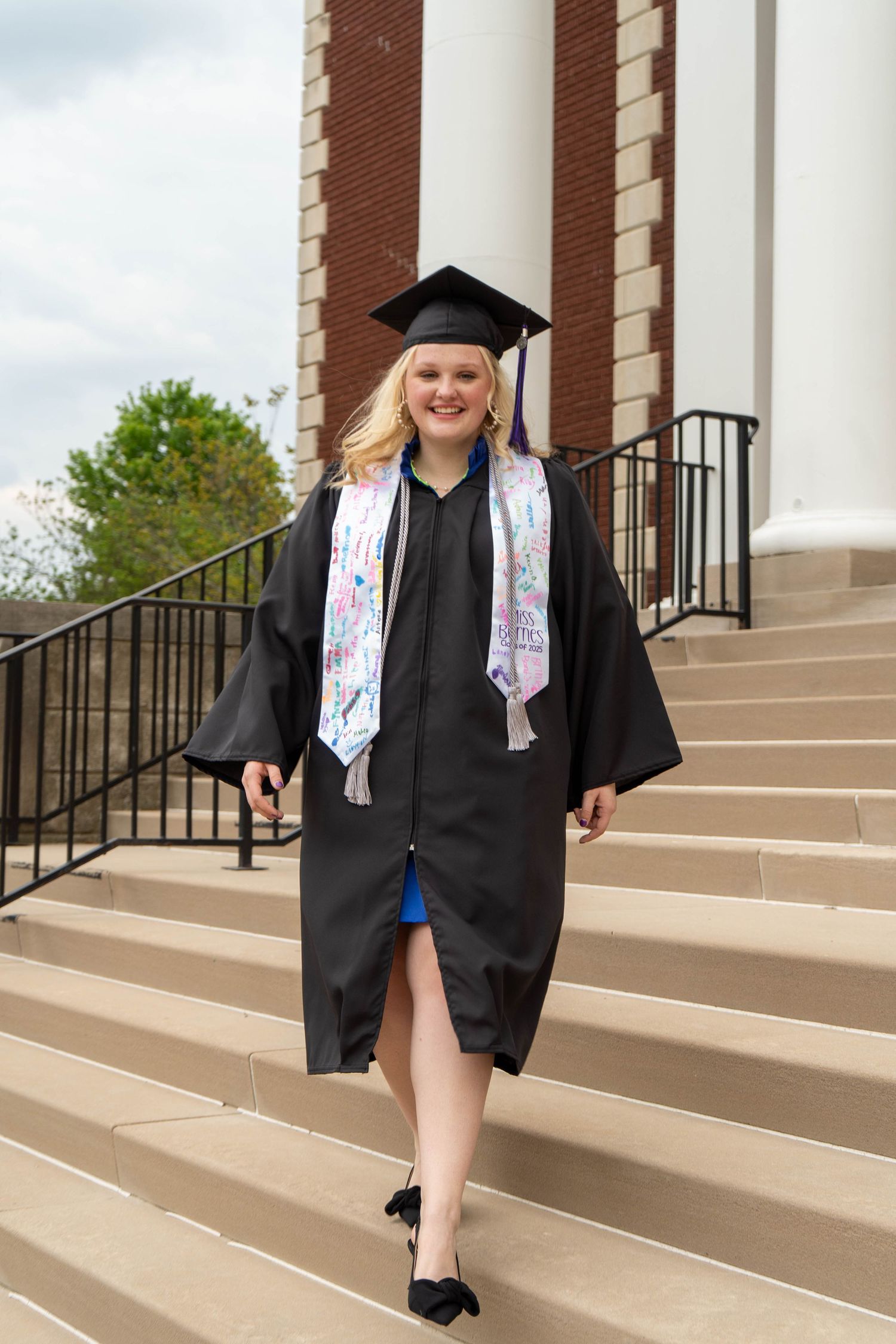 Blonde graduate in black cap and gown walking down university building steps.