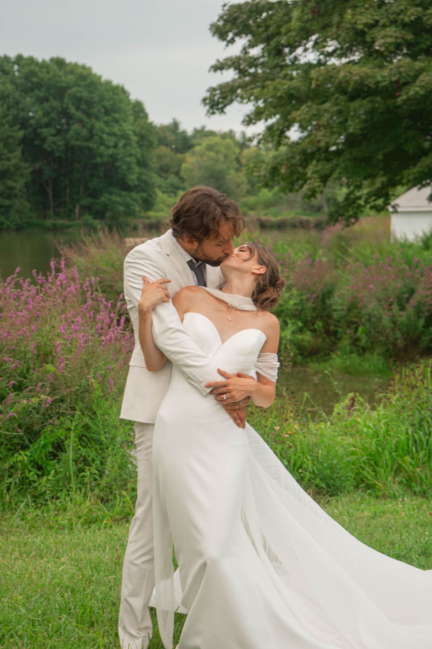 Groom in cream suit dips and kisses bride in white off-shoulder gown beside a wildflower-lined pond.