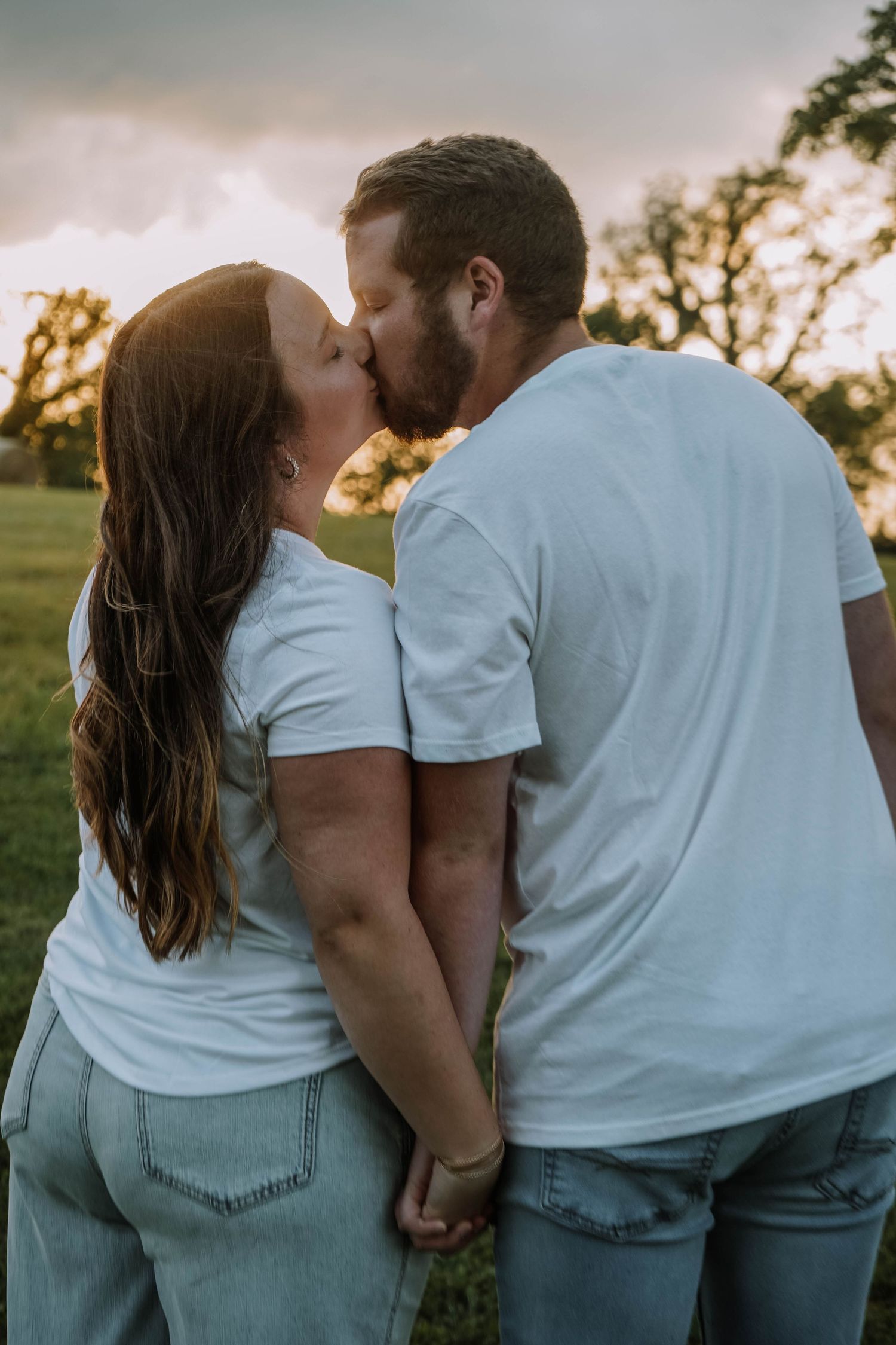 Couple in white tees kissing while holding hands in a green field during a dramatic sunset.