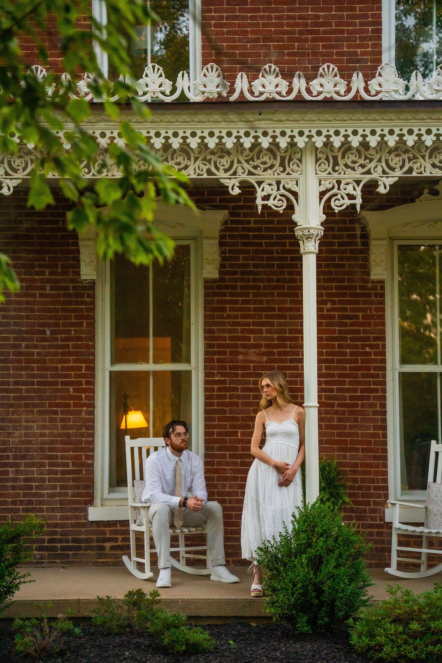 Couple on Victorian brick house porch, woman in white dress standing as man sits in rocking chair.