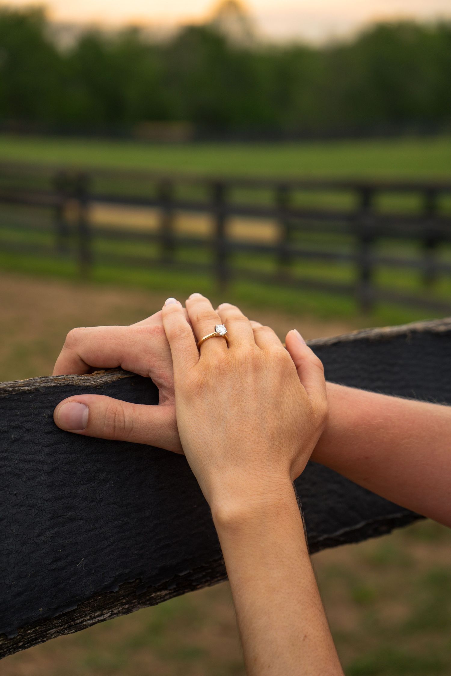 Close-up of couple's hands on black fence rail showing diamond engagement ring at sunset.