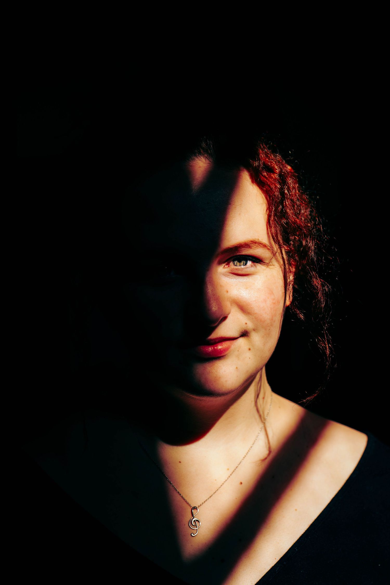 Redhead woman smiling with dramatic split lighting, wearing a treble clef necklace against a black background.