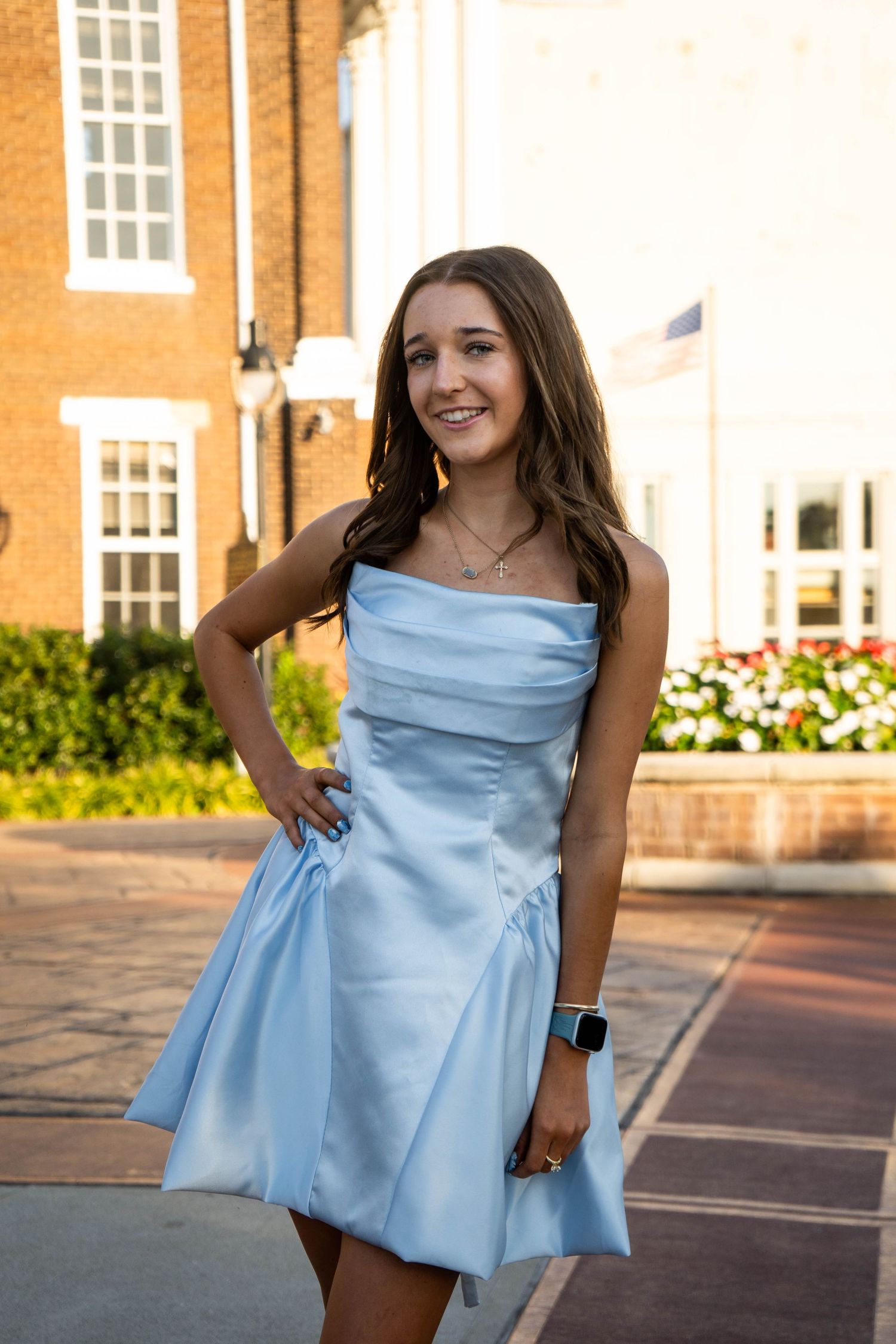 Smiling young woman in light blue strapless satin dress poses outdoors near brick building.
