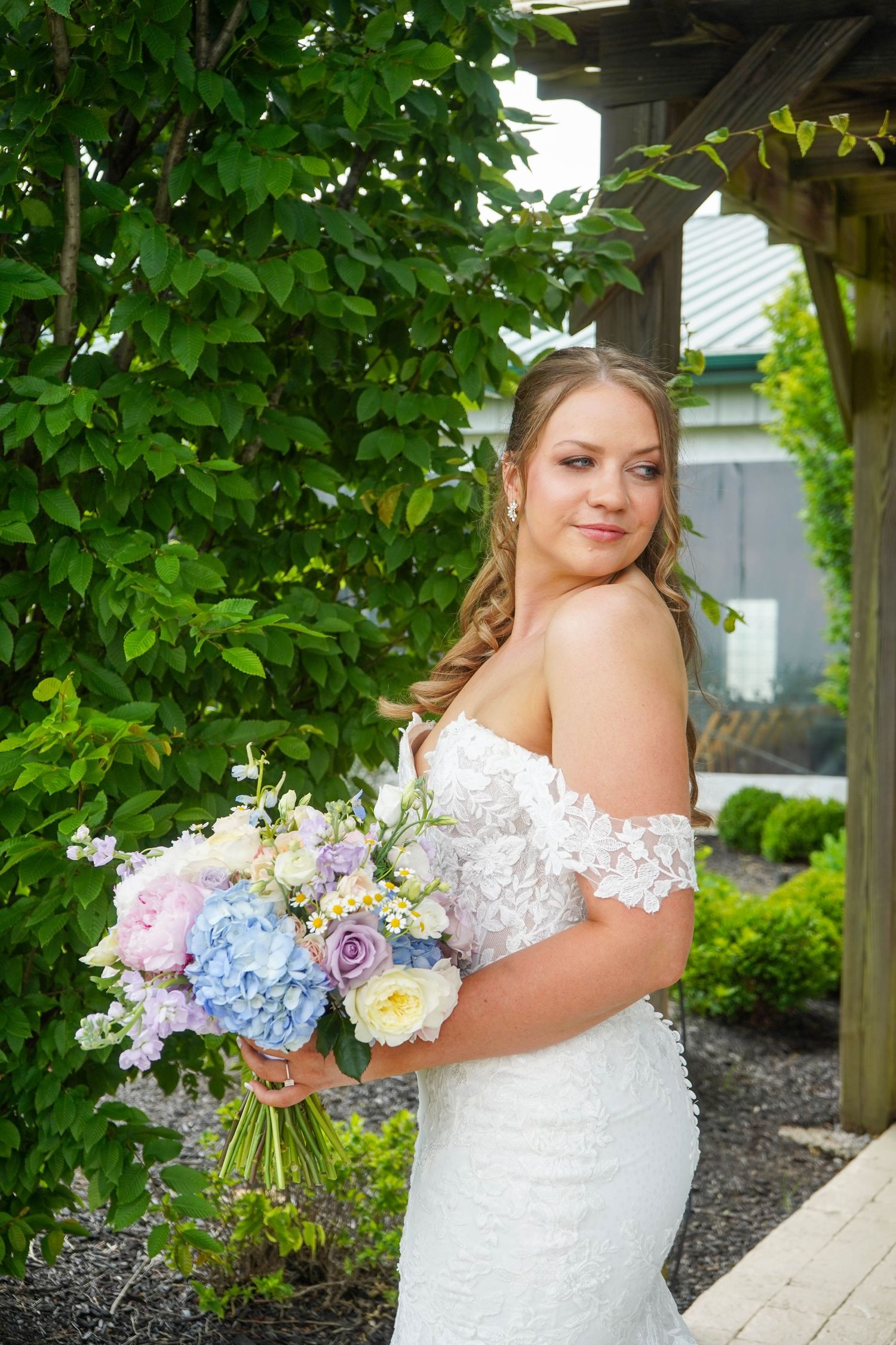 Bride in off-shoulder lace wedding gown holds pastel blue and pink hydrangea bouquet in garden setting.