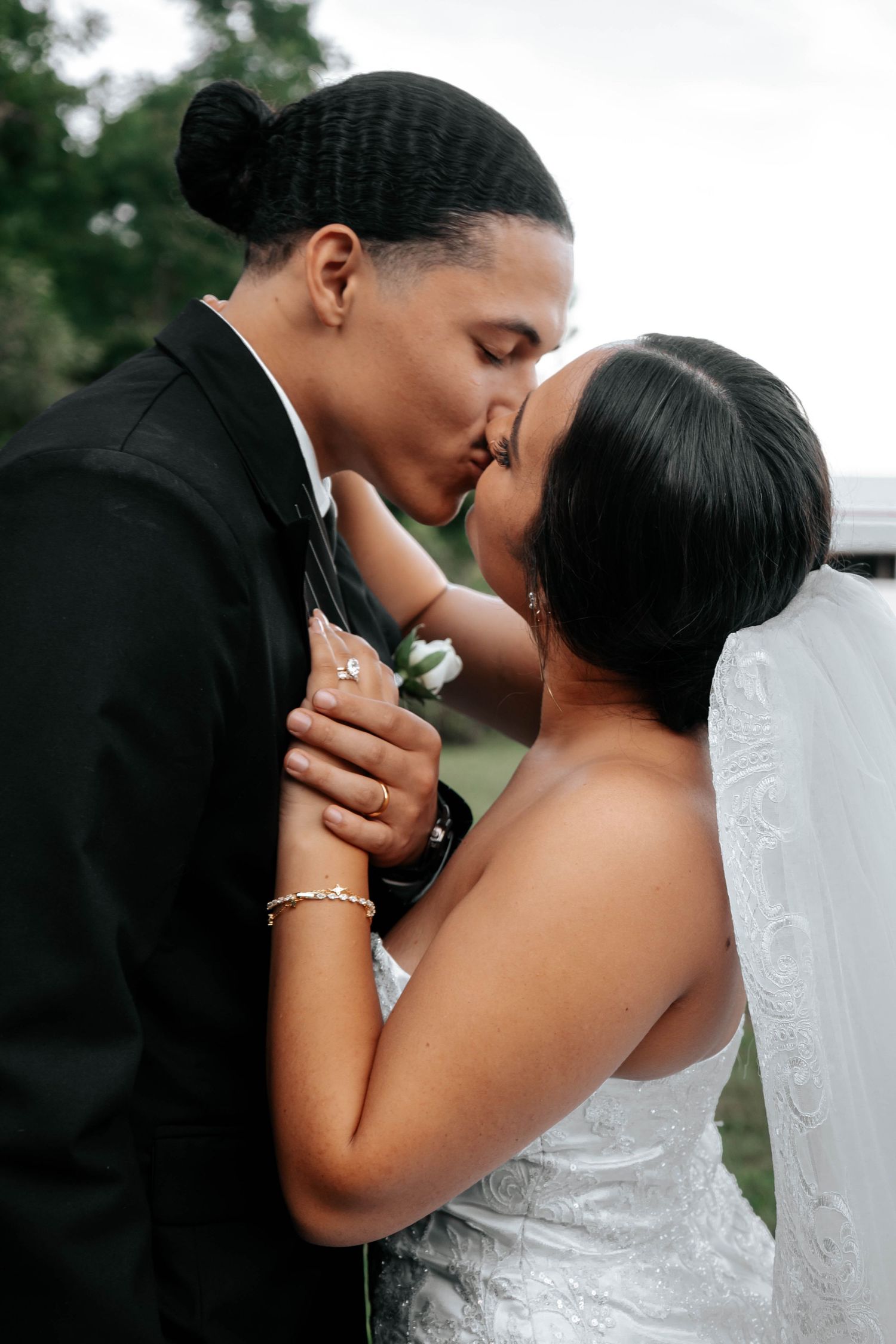 Bride and groom share a romantic kiss outdoors on their wedding day, with bride in lace gown and veil.