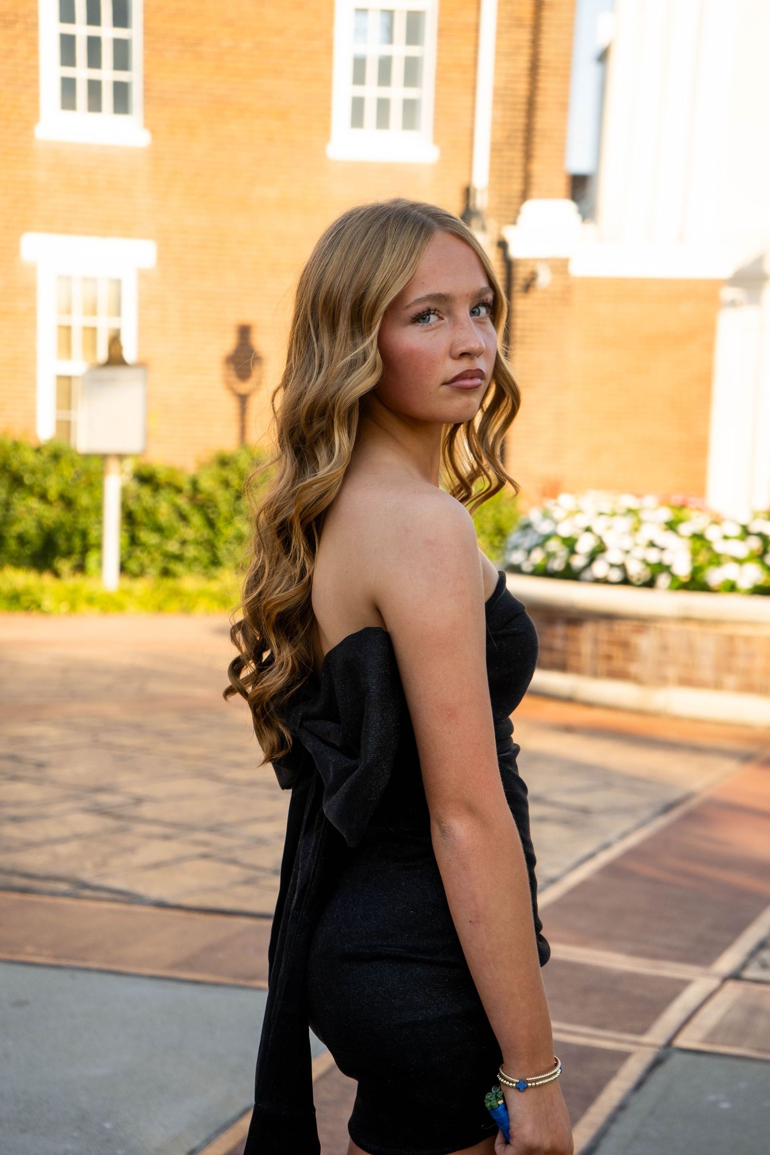 Blonde teenage girl in strapless black bow-back dress poses outdoors near brick building at sunset.