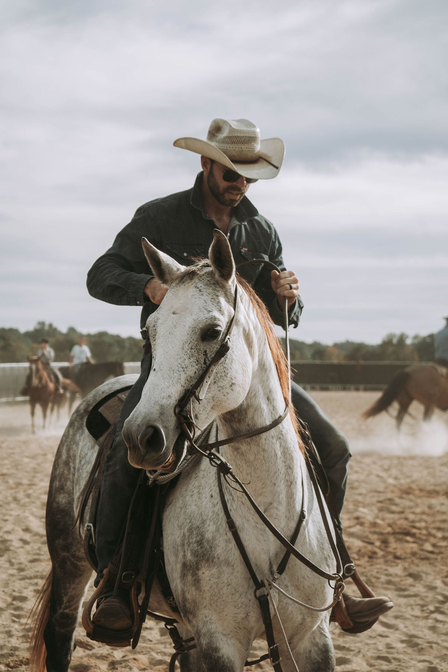 Cowboy in white hat riding gray horse in sandy arena with other horses visible in background.