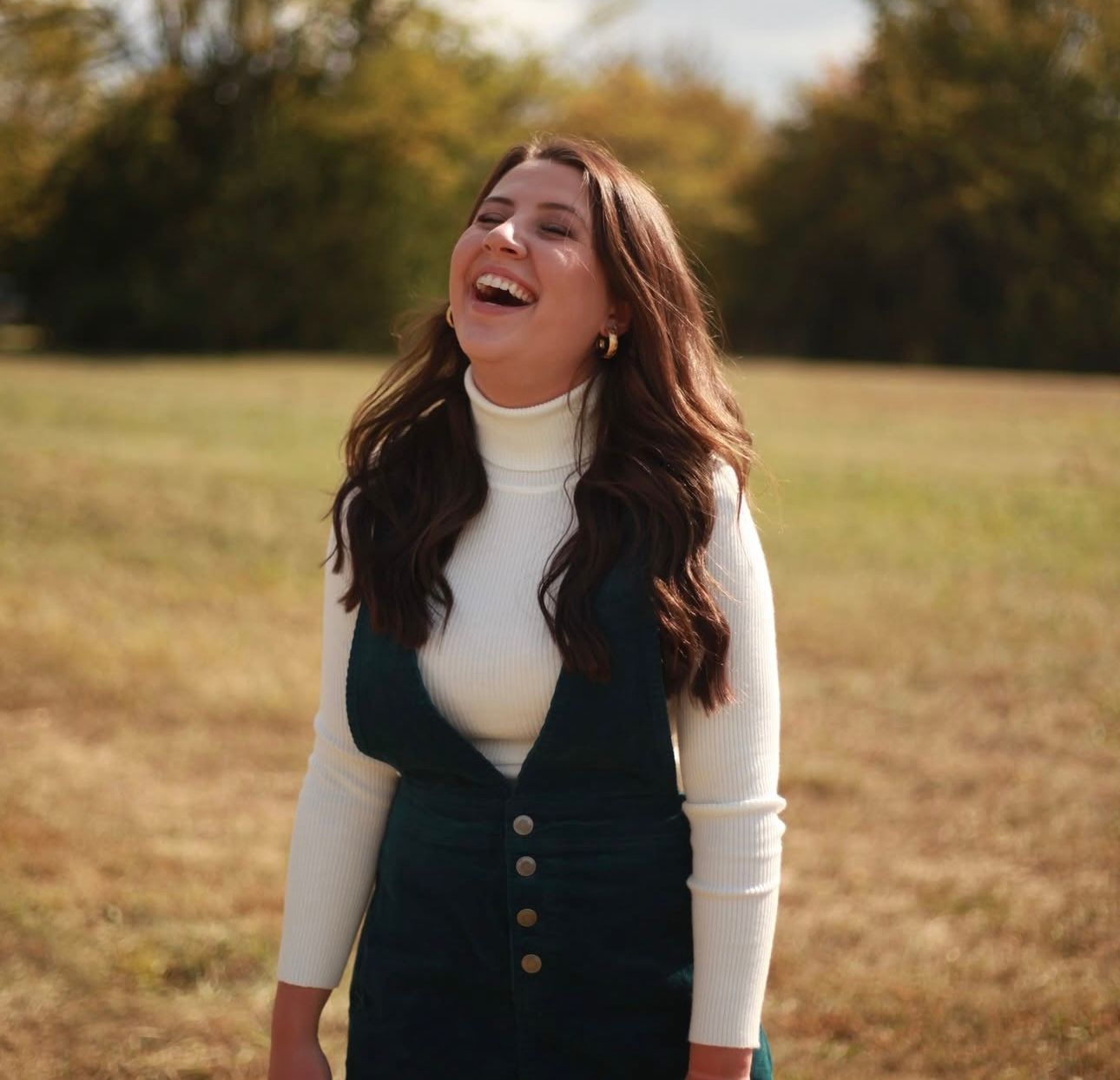 Young woman laughing joyfully in an open field wearing a white turtleneck and dark jumper.