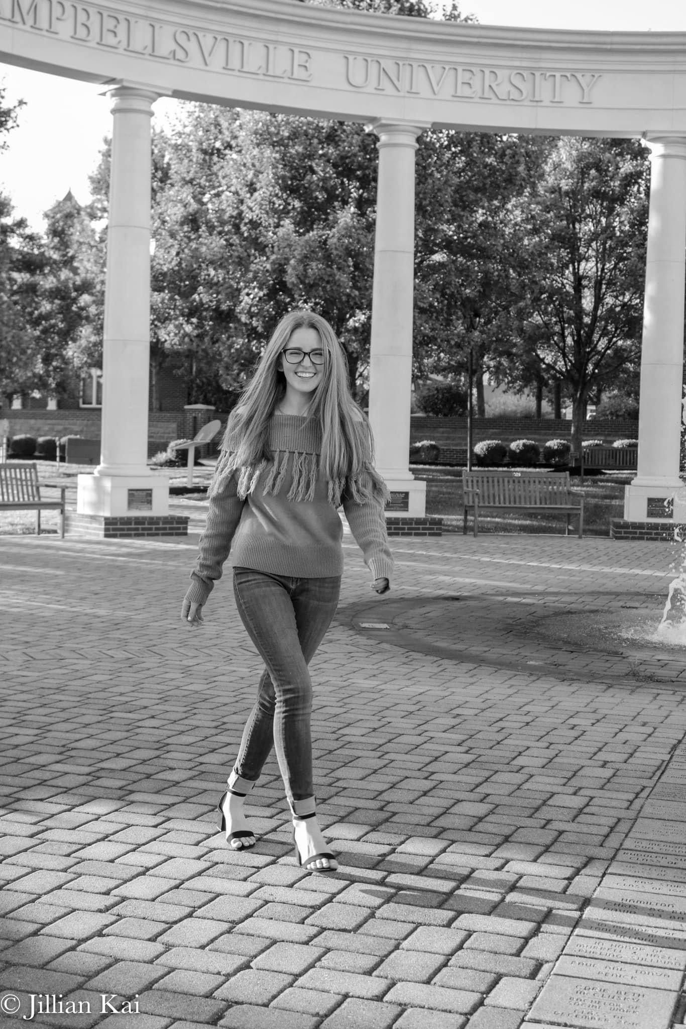 Smiling young woman walks on brick path near Campbellsville University arch in black and white.