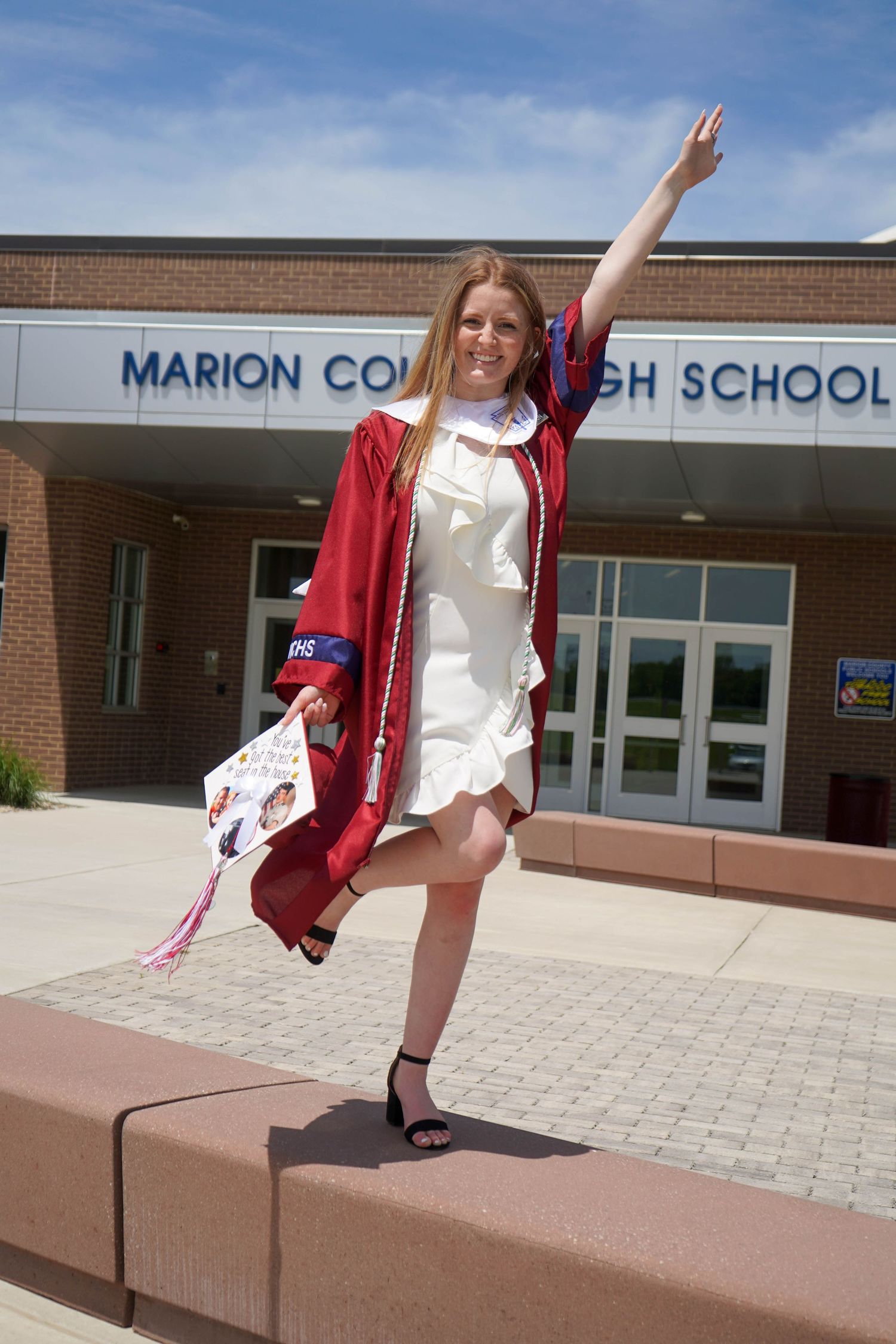 Smiling graduate in red gown celebrates outside Marion County High School, raising hand joyfully.